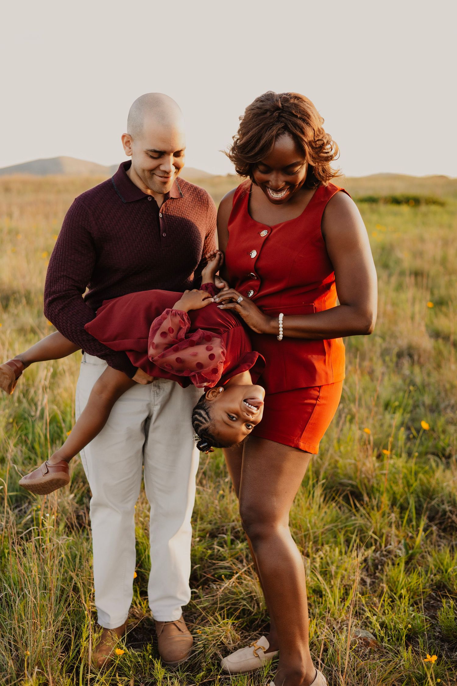 A joyful family plays in a green field at sunset, with parents laughing as they flip their smiling child upside down.