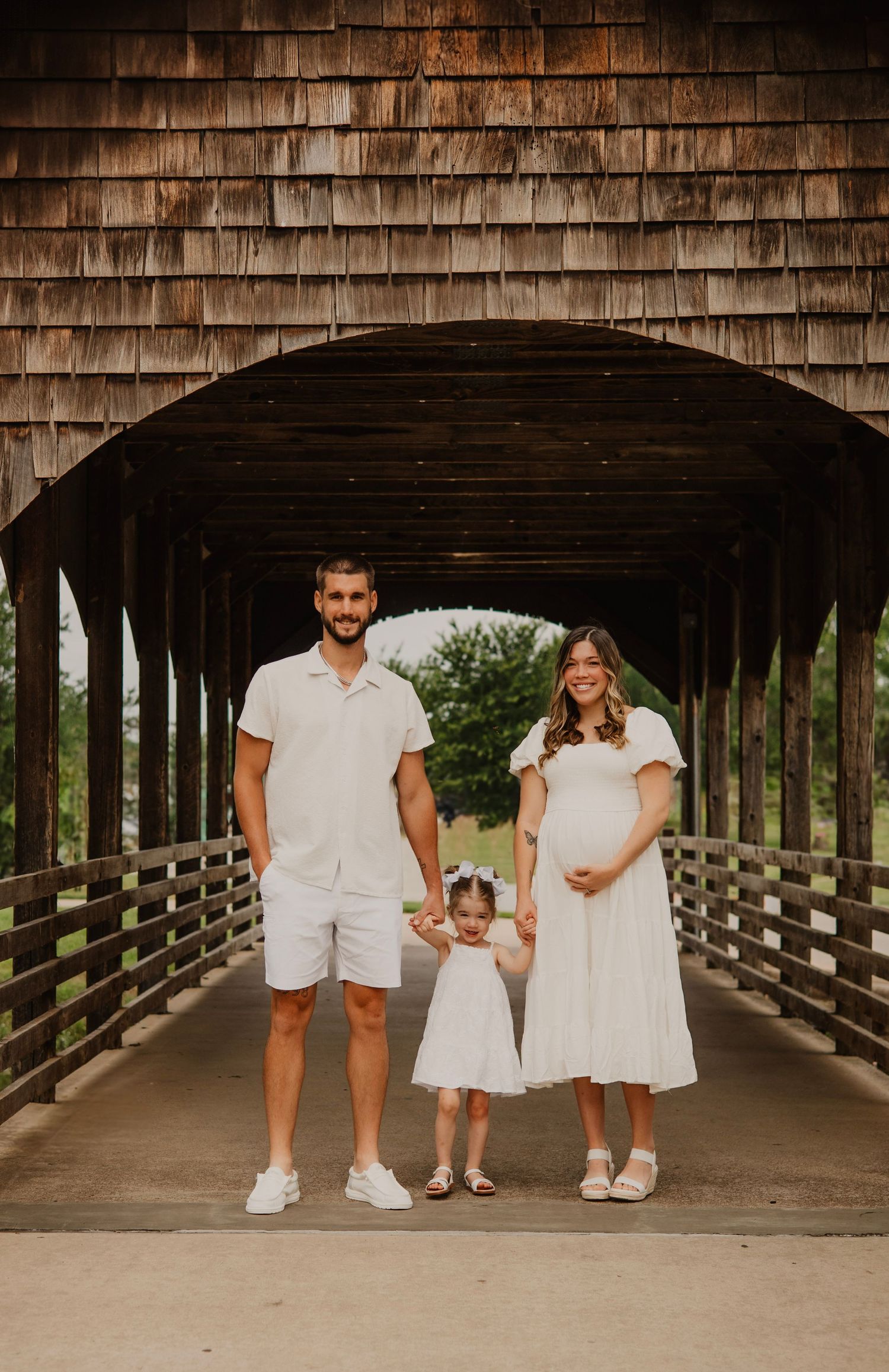 A family in coordinated white outfits poses together on a covered wooden bridge during a summer photo session.