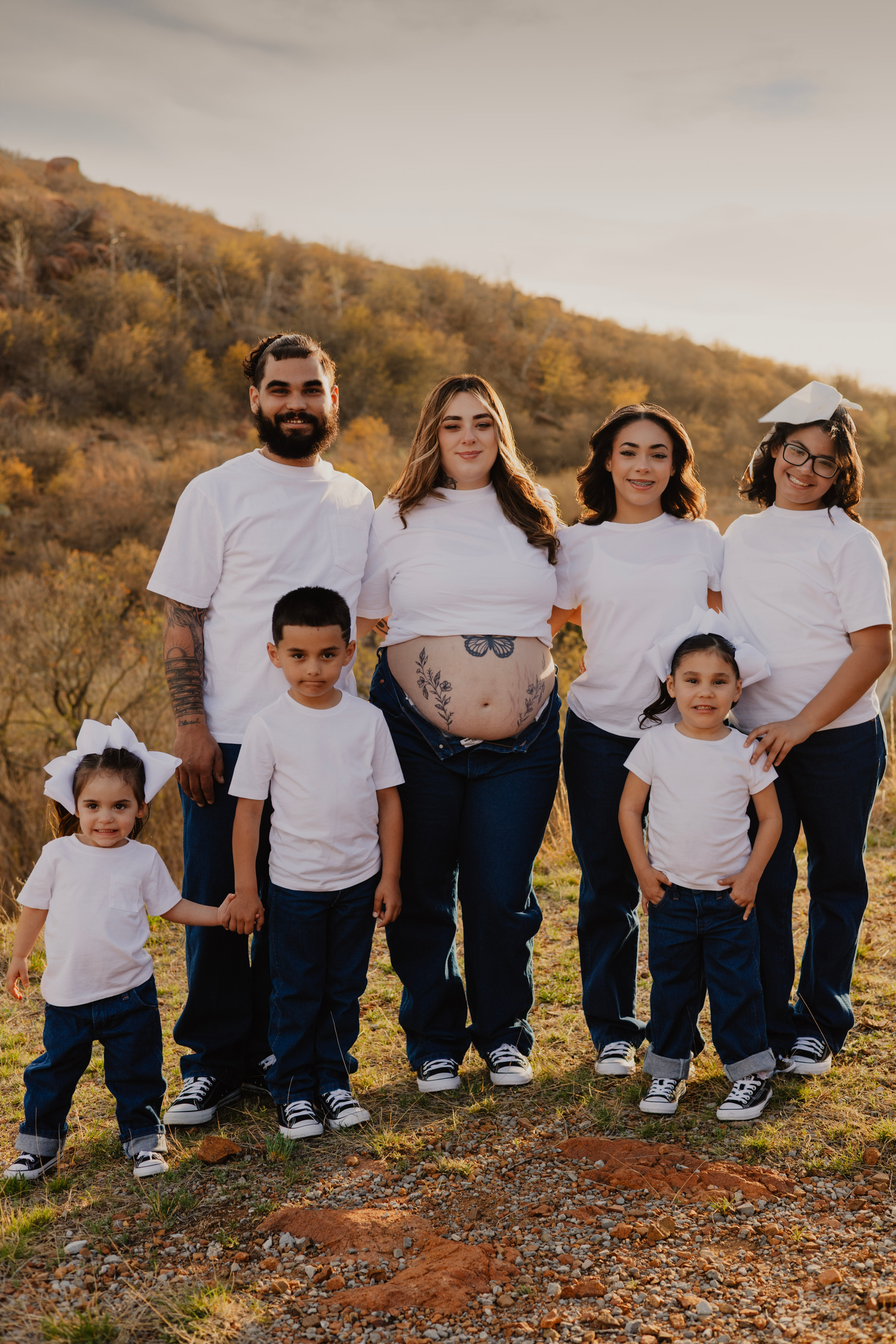 Family of seven in matching white tees and jeans poses outdoors, with pregnant mother showing baby bump tattoo.