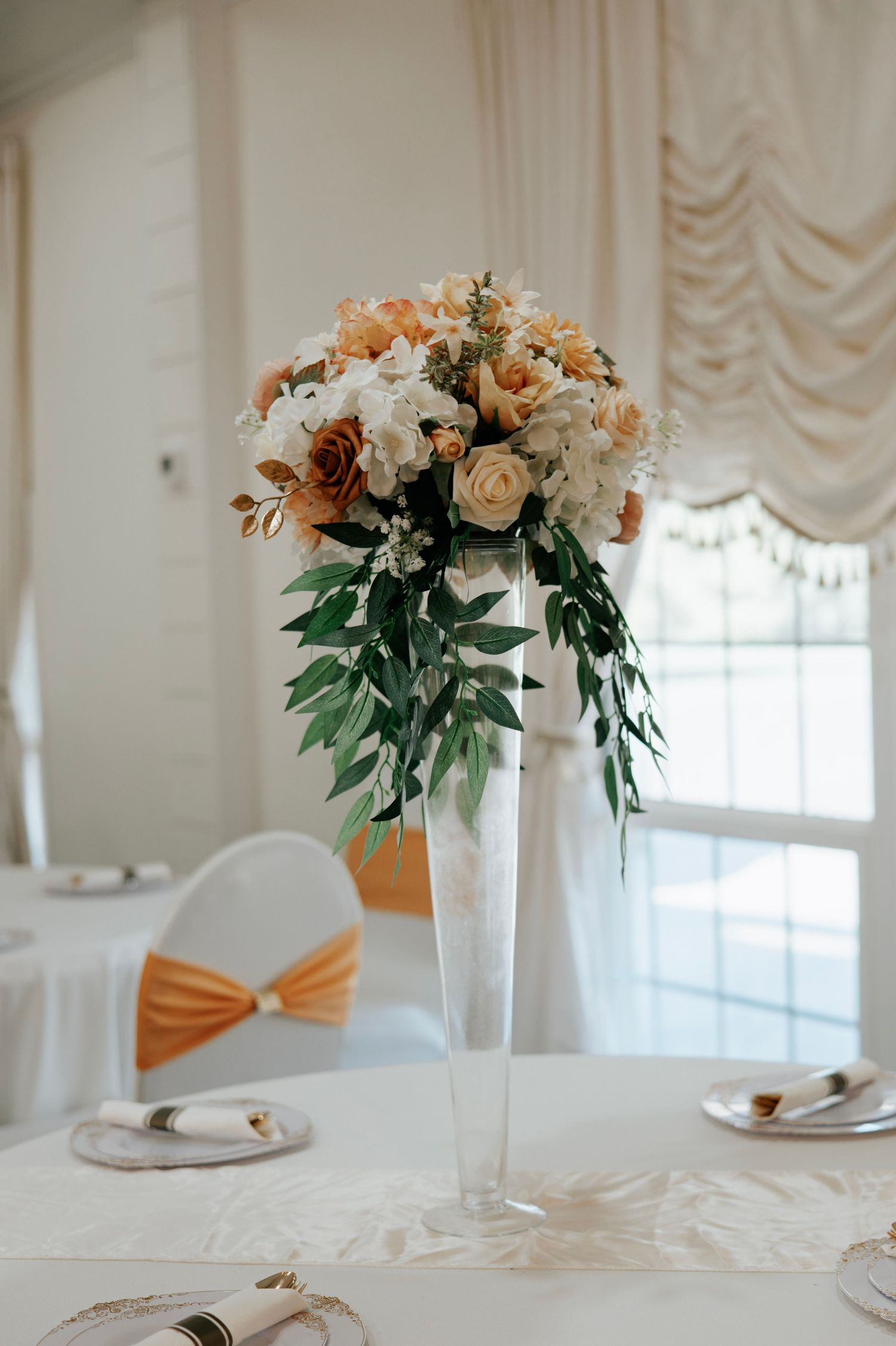 Elegant wedding reception table with tall white floral centerpiece and peach accents in a bright banquet room.