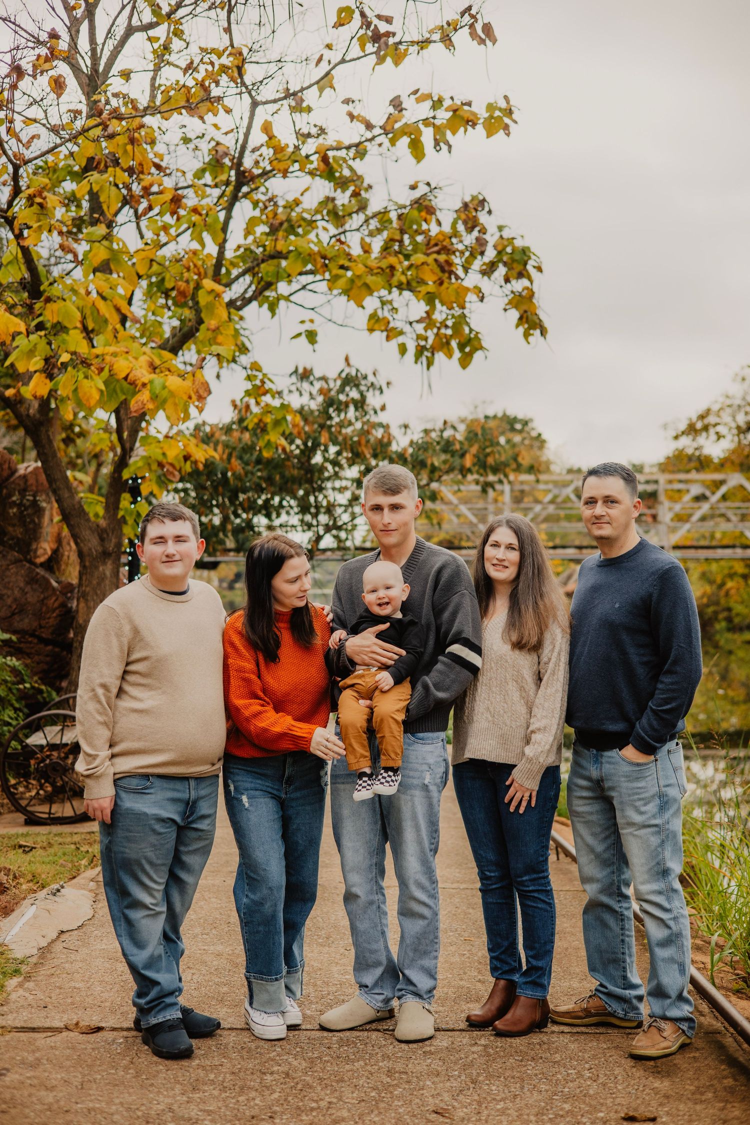 Family group poses together on path with autumn trees wearing coordinated neutral tones.