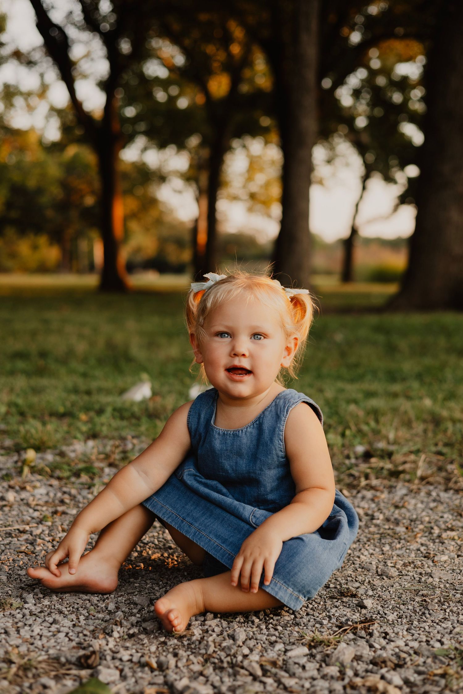 Young child in denim dress sits barefoot on gravel path with trees in background.