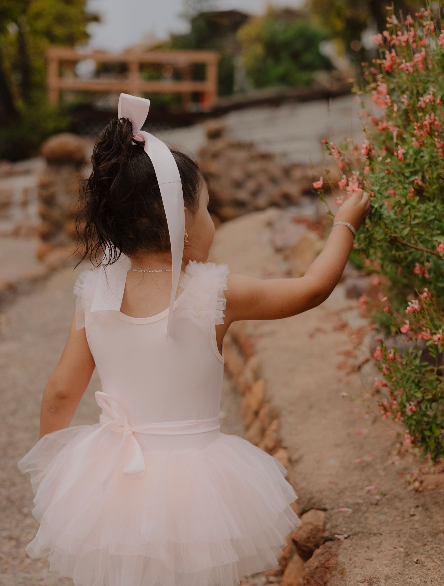 A small child in a white tutu dress reaches toward pink flowers on a garden path.