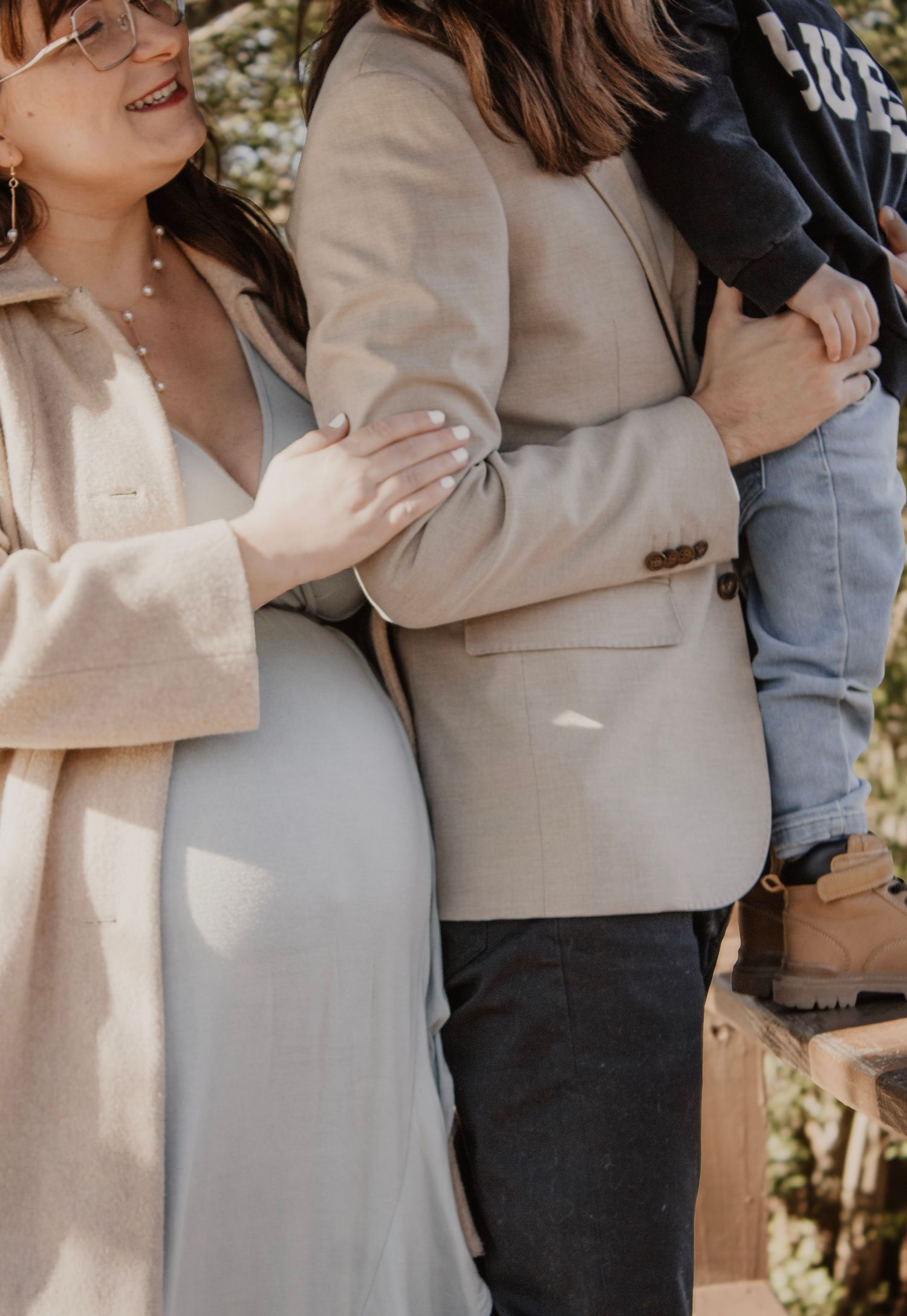 Three people embrace while showing off baby bumps in neutral-toned clothing.
