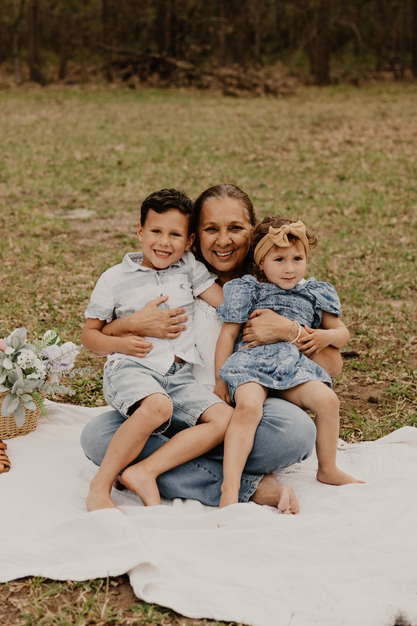 Grandmother sitting on white blanket outdoors, hugging young boy and toddler girl in a grassy field.