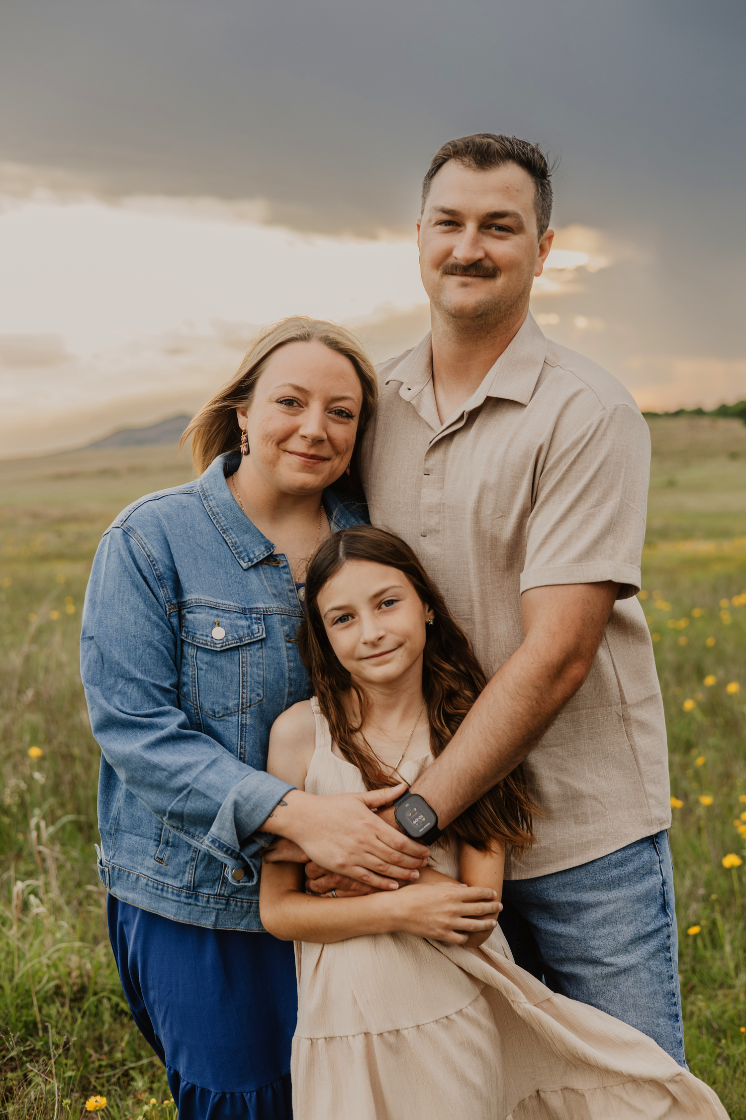Family of three posing in a green meadow with wildflowers and dramatic cloudy sky at golden hour.