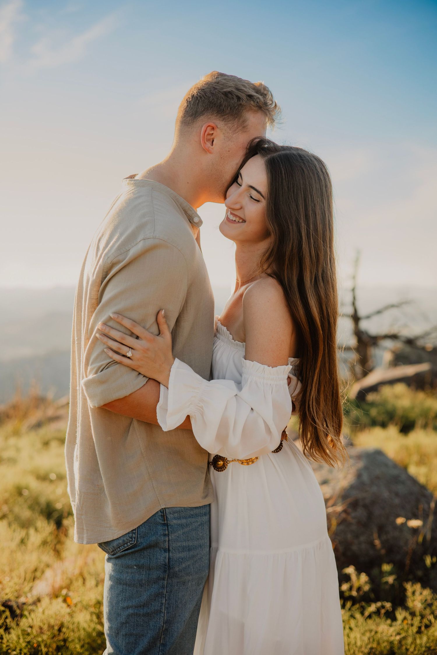 A tender embrace between a couple in white and beige clothing against a mountain landscape.