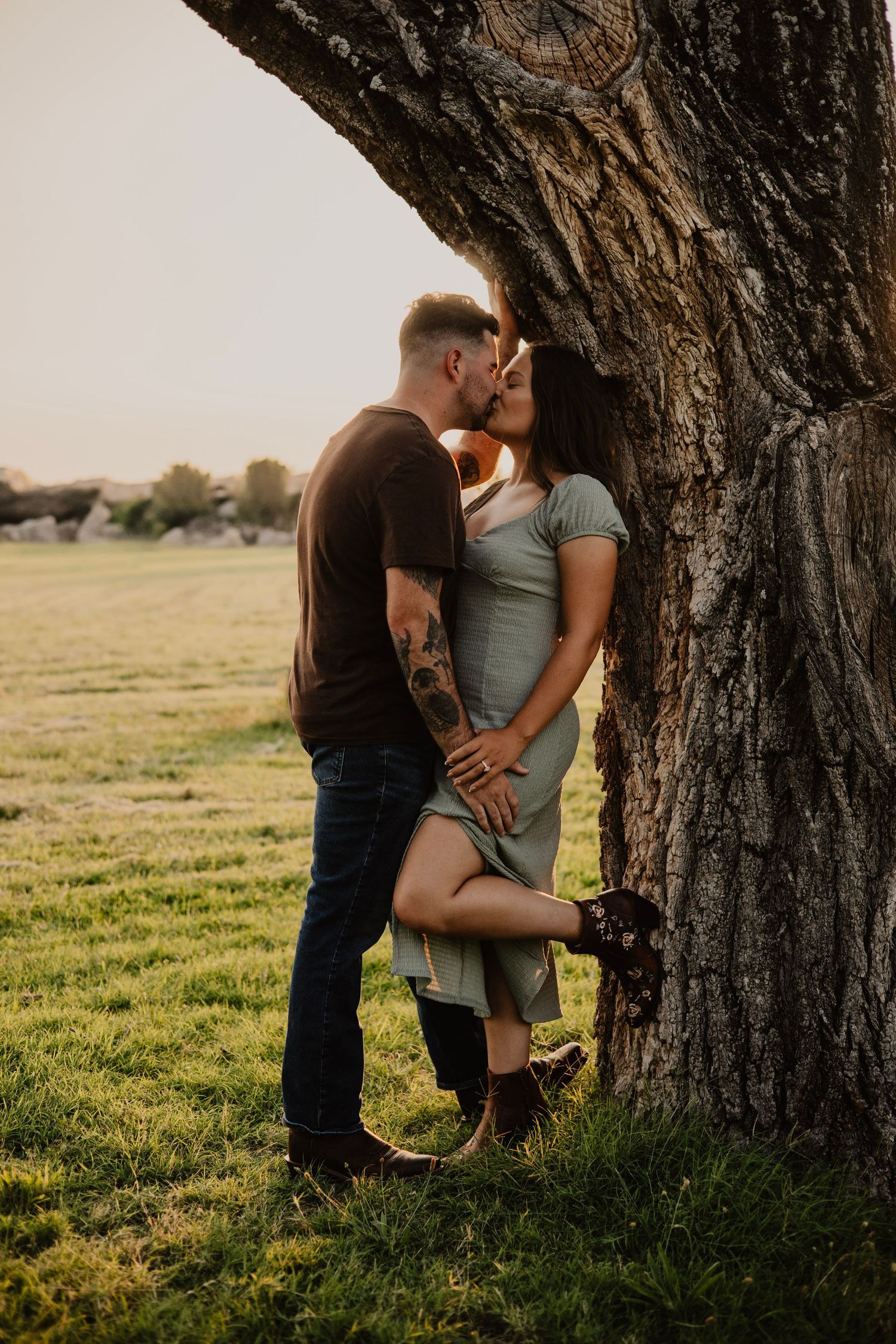 Romantic couple embracing against large tree trunk at sunset in a grassy field.