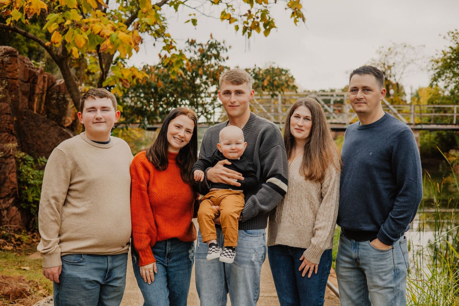 Group photo of family members in casual sweaters standing by autumn pond.