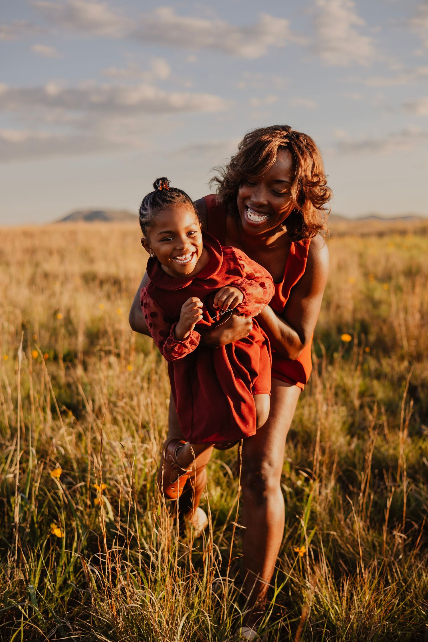 A joyful woman gives a laughing young girl a piggyback ride in a golden sunlit field, both wearing red dresses.
