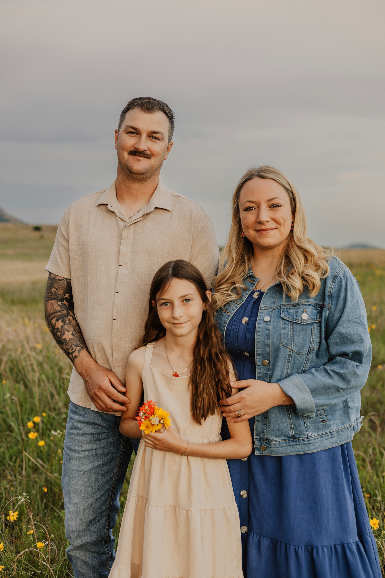 A family of three poses in a wildflower field, with a girl holding orange flowers between her tattooed father and smiling mother.