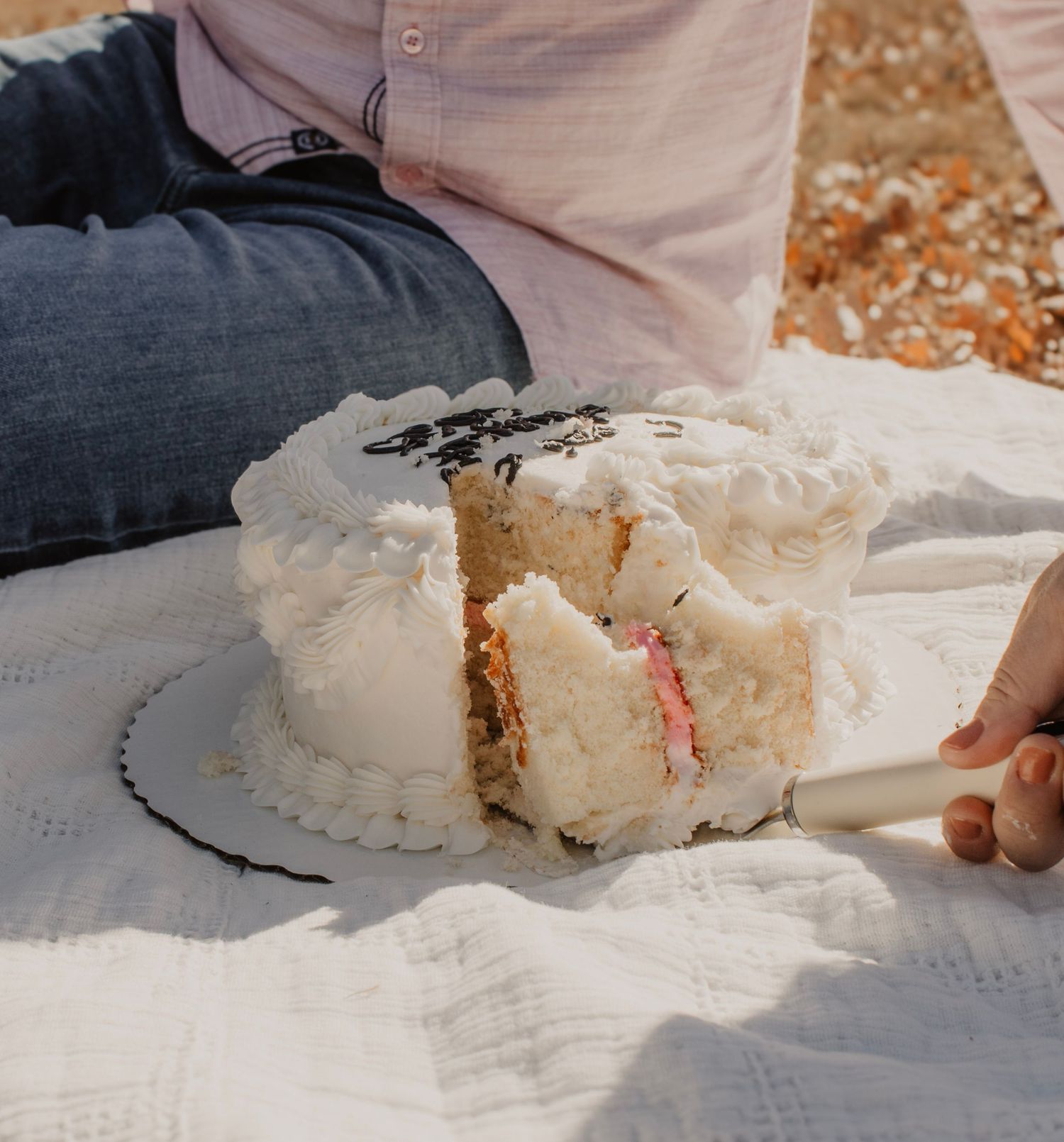 A white frosted cake being cut on a picnic blanket outdoors.