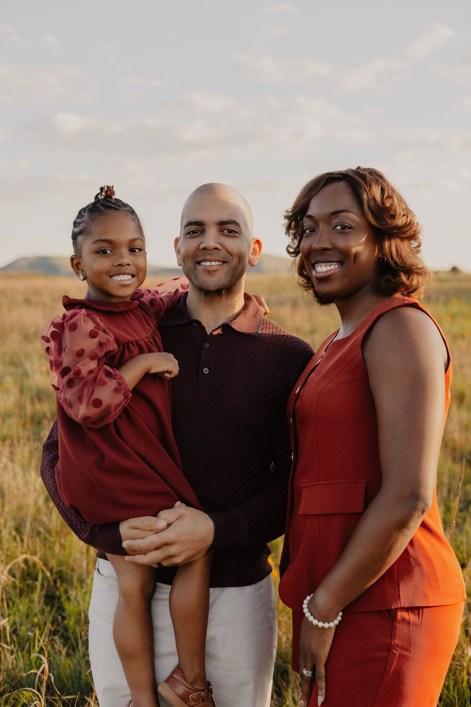 A smiling family of three poses outdoors in a field, with a man holding a young girl and a woman beside them.