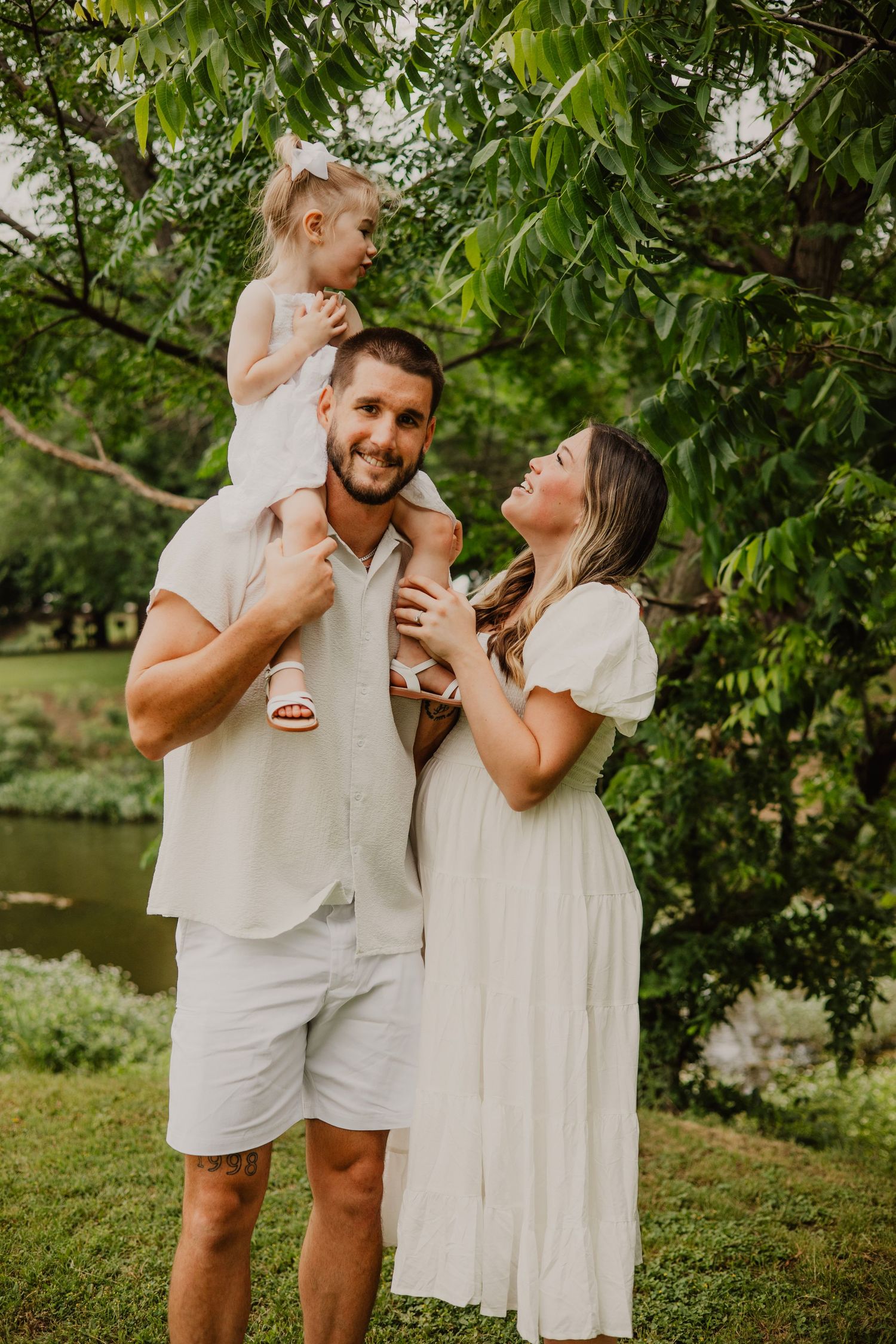 Family in white outfits enjoying a summer day together by a pond under green leafy trees.