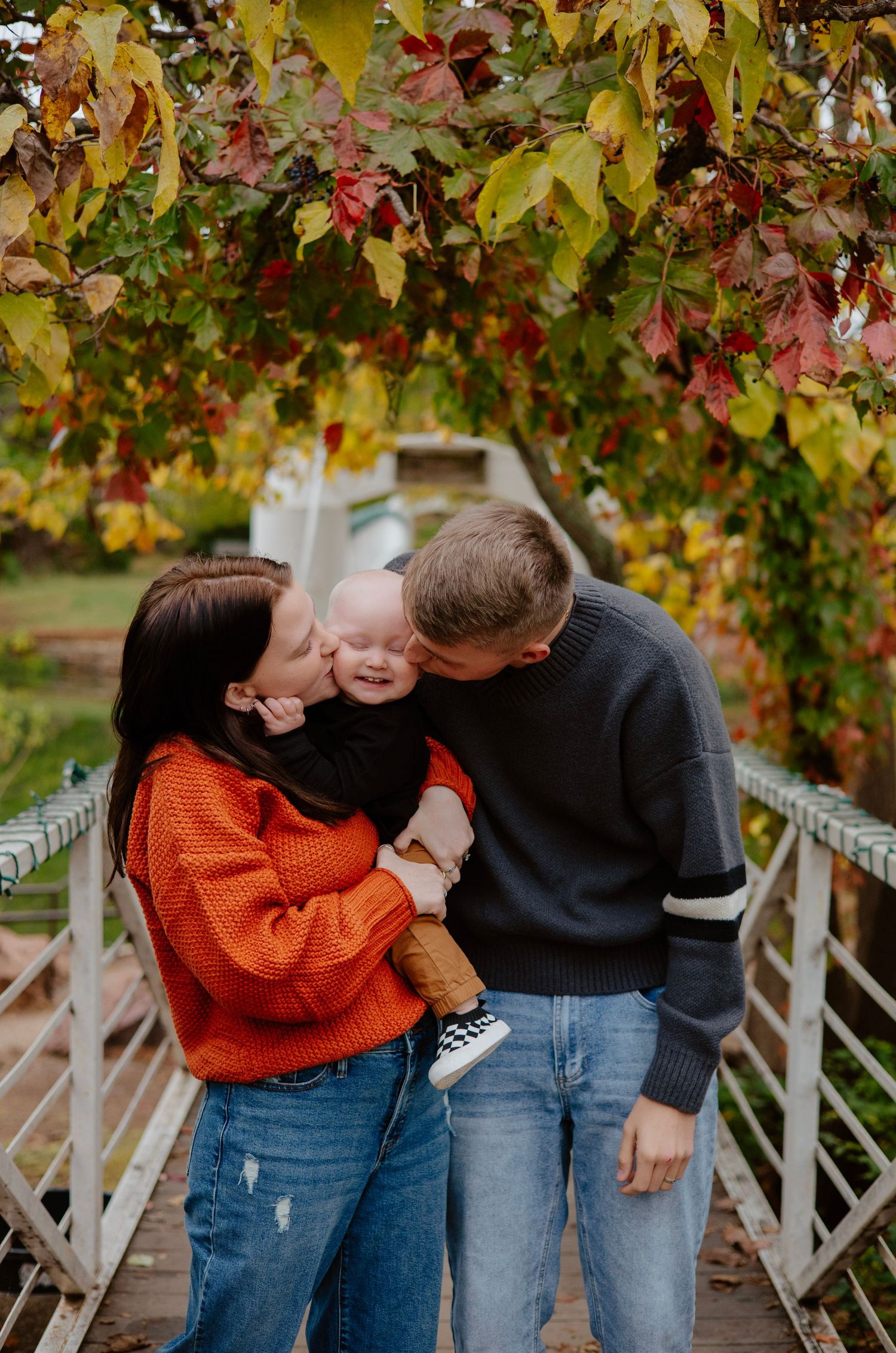 Family shares tender moments together under autumn foliage with orange sweater and dark clothing.