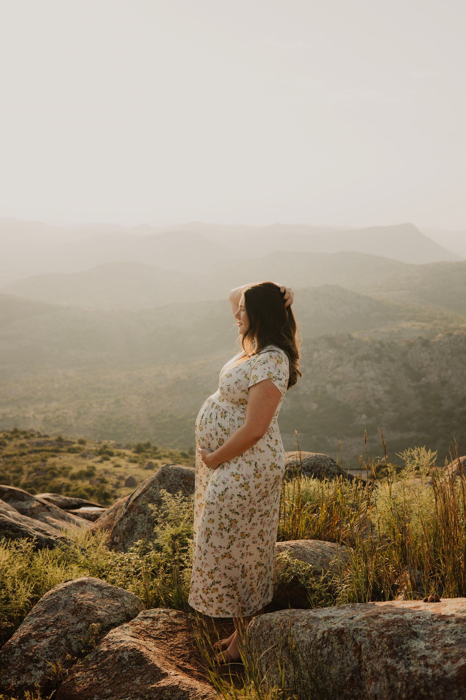 Silhouette of an expectant mother in a floral dress standing on rocks overlooking mountains at sunset.