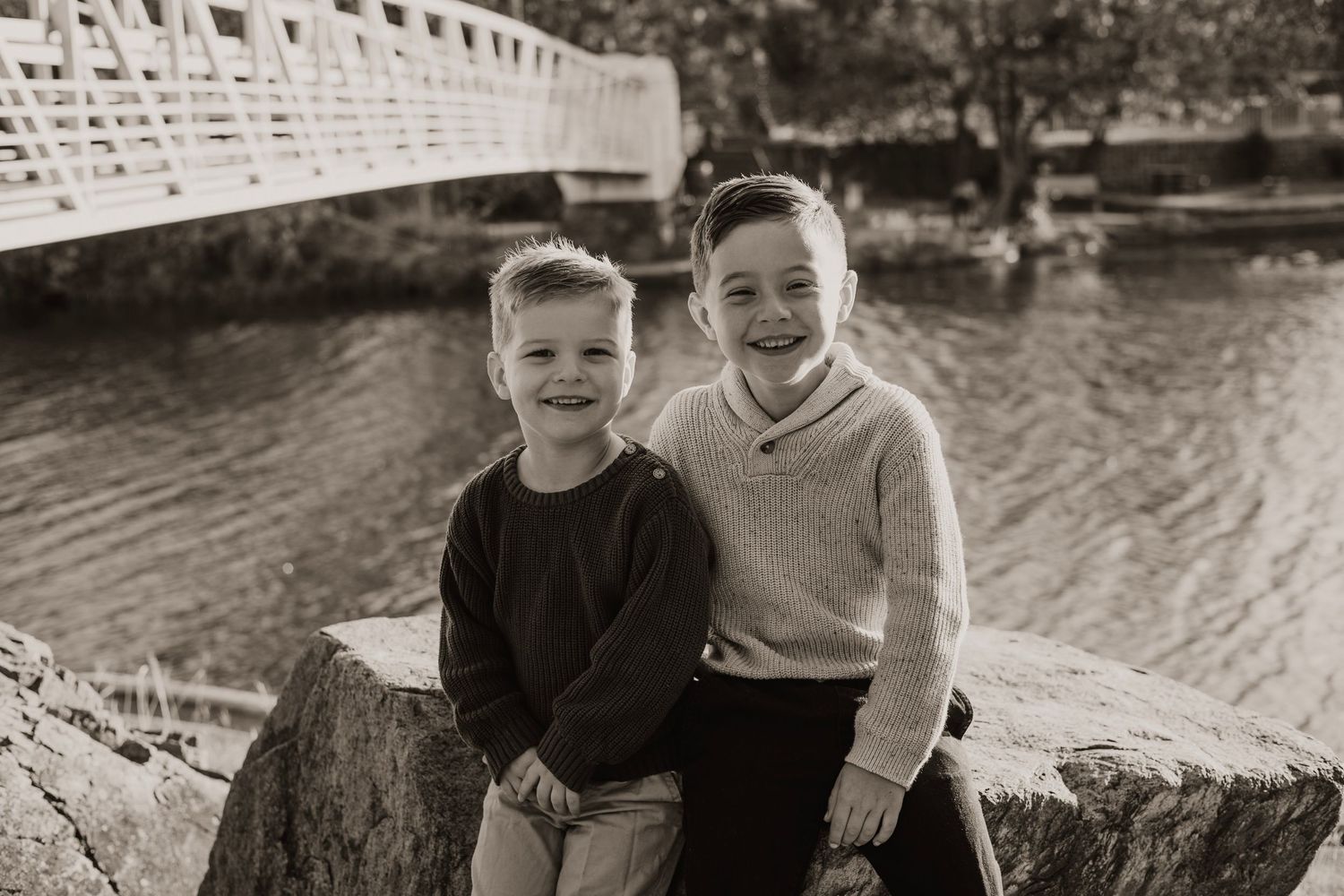 Two children sit together on log by river with white bridge in background.