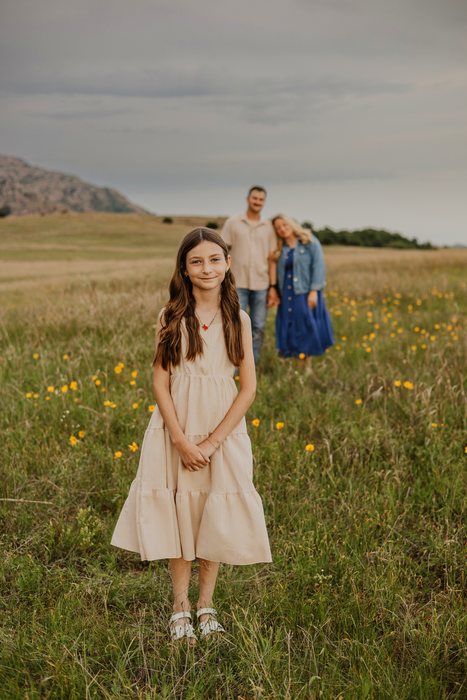 A young girl in a beige dress stands in a flower field with her parents blurred in the background near mountains.