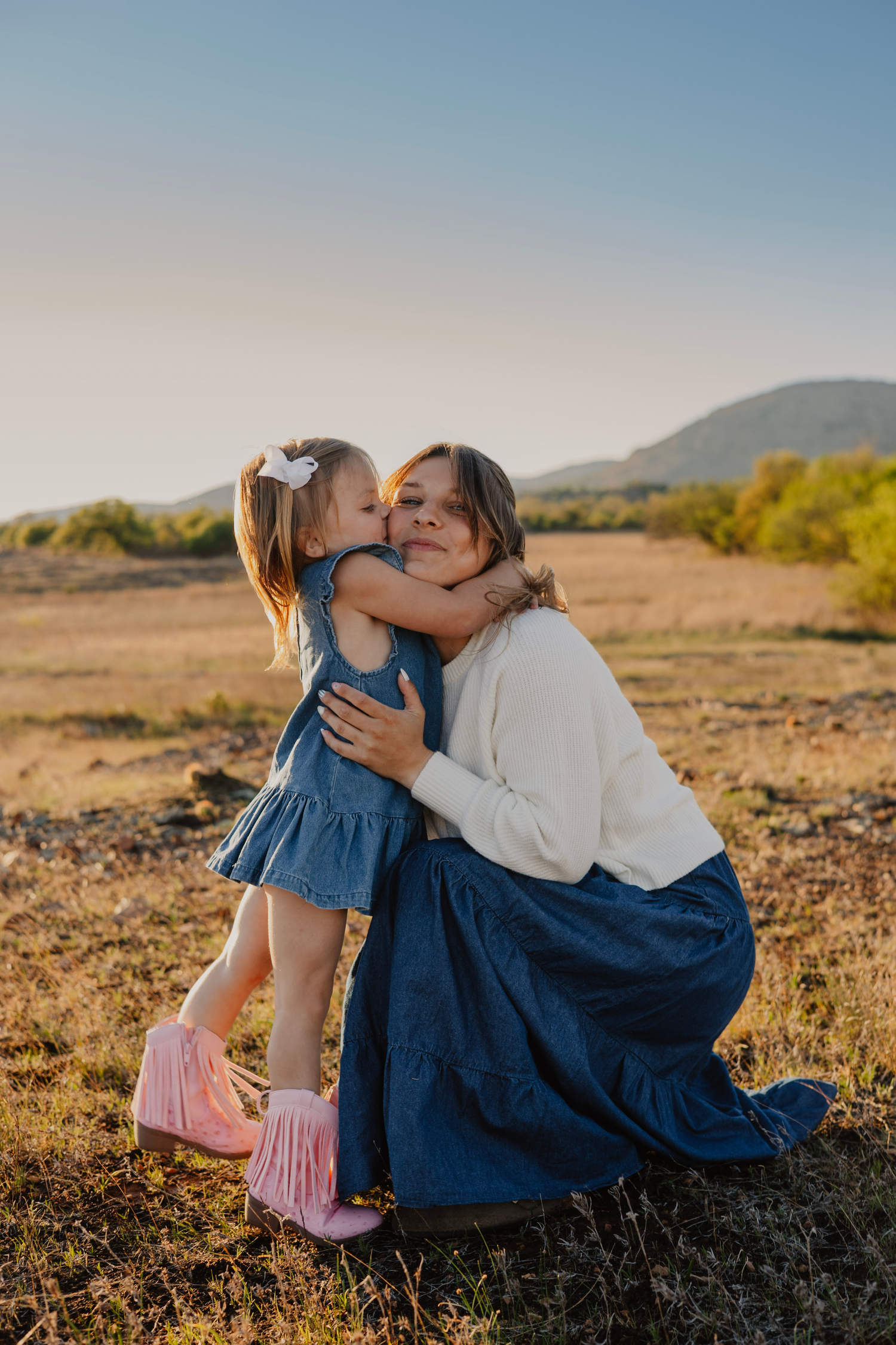 A mother kneels in a golden field as her young daughter in a blue dress and pink boots gives her a sweet kiss.