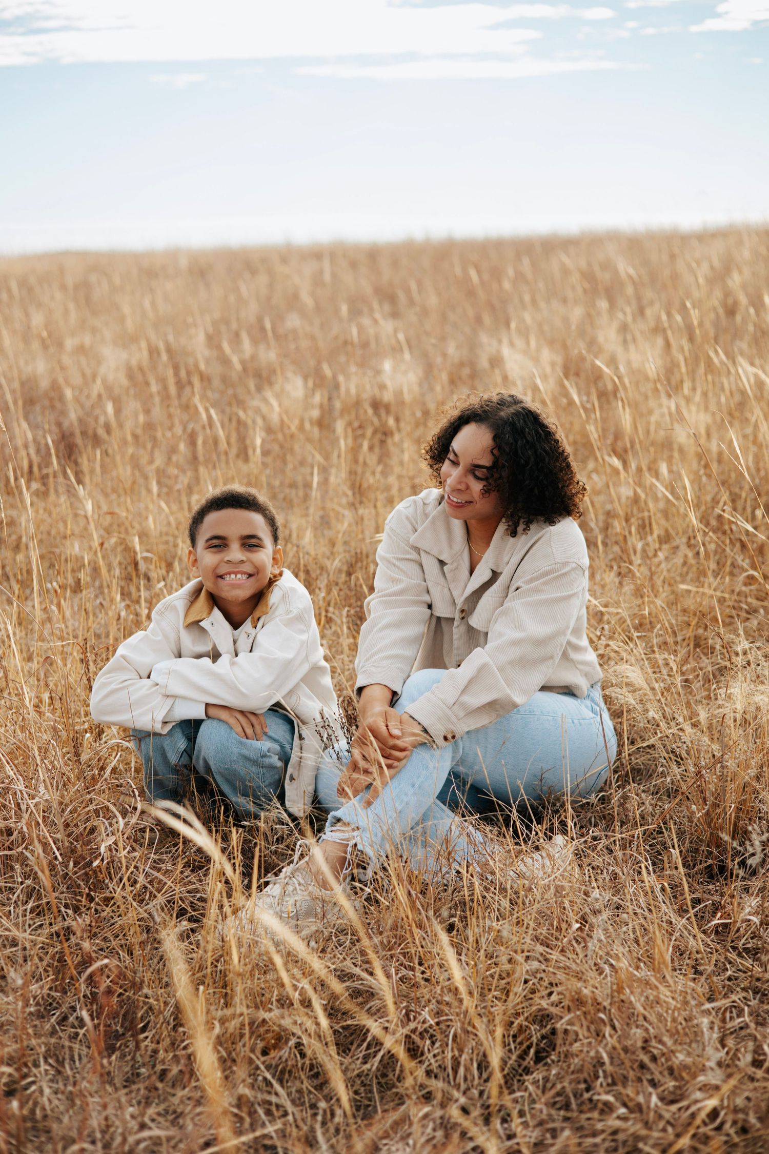 Two people in white tops and jeans sit together in a golden wheat field on a sunny day.
