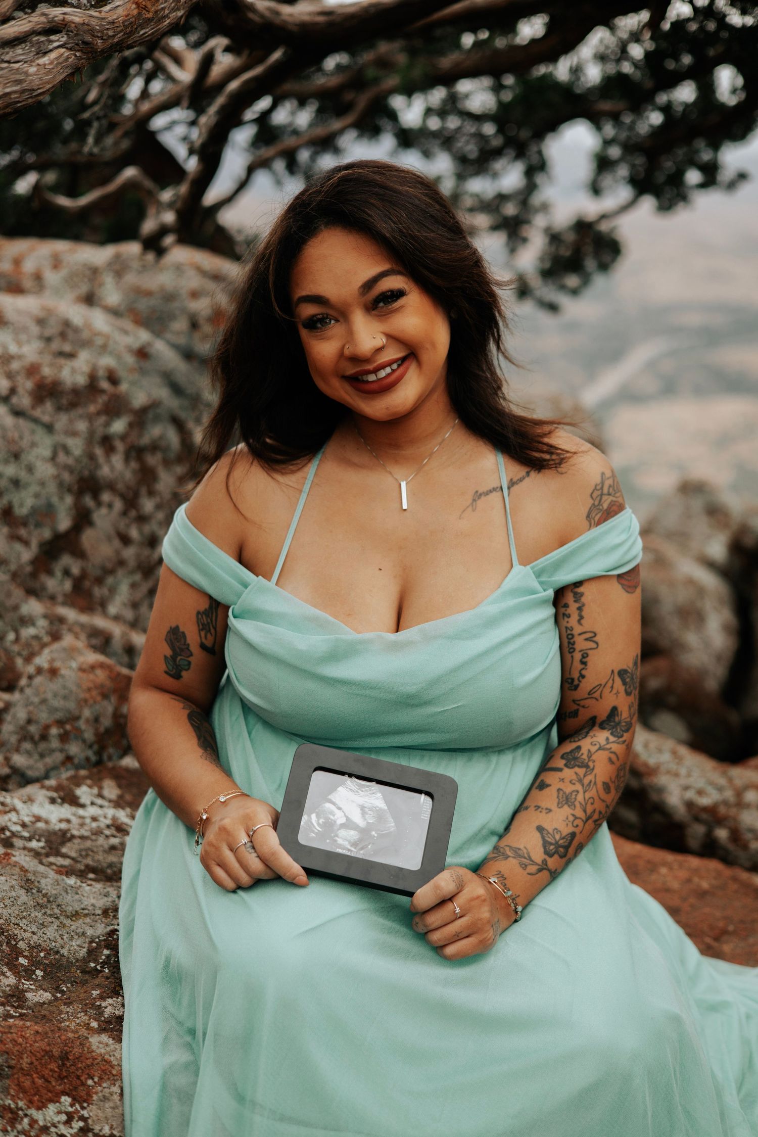 A woman in a mint green dress poses outdoors holding an ultrasound photo against a mountain backdrop.