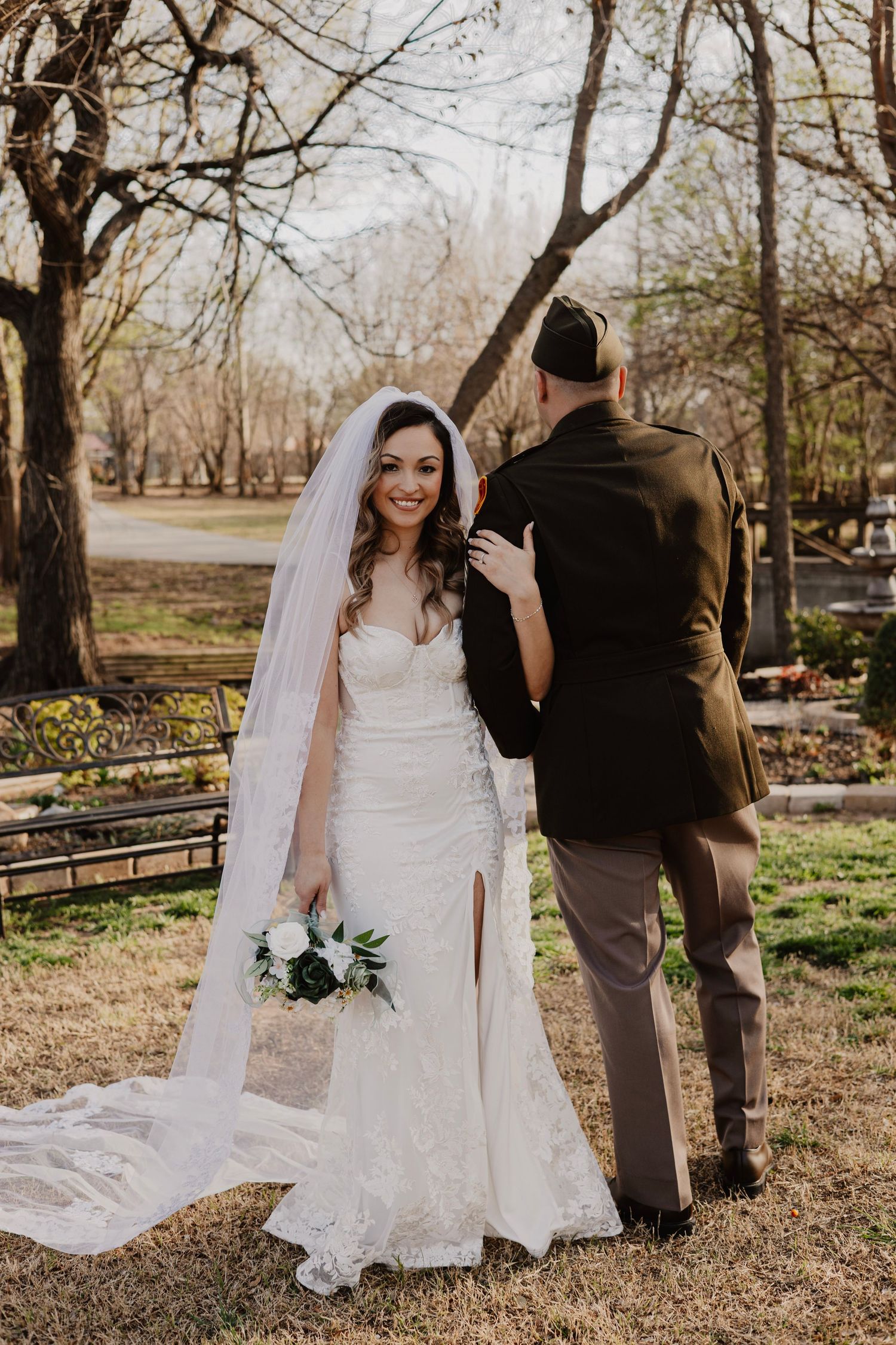 A bride in a lace wedding dress poses outdoors in a park setting with trees and grass on a sunny day.