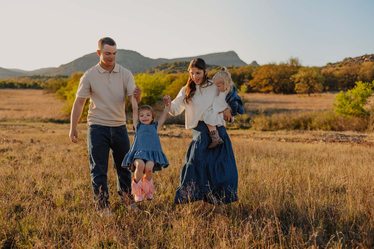 Family of four walking in golden field with mountains, parents swinging young daughter in blue dress at sunset.