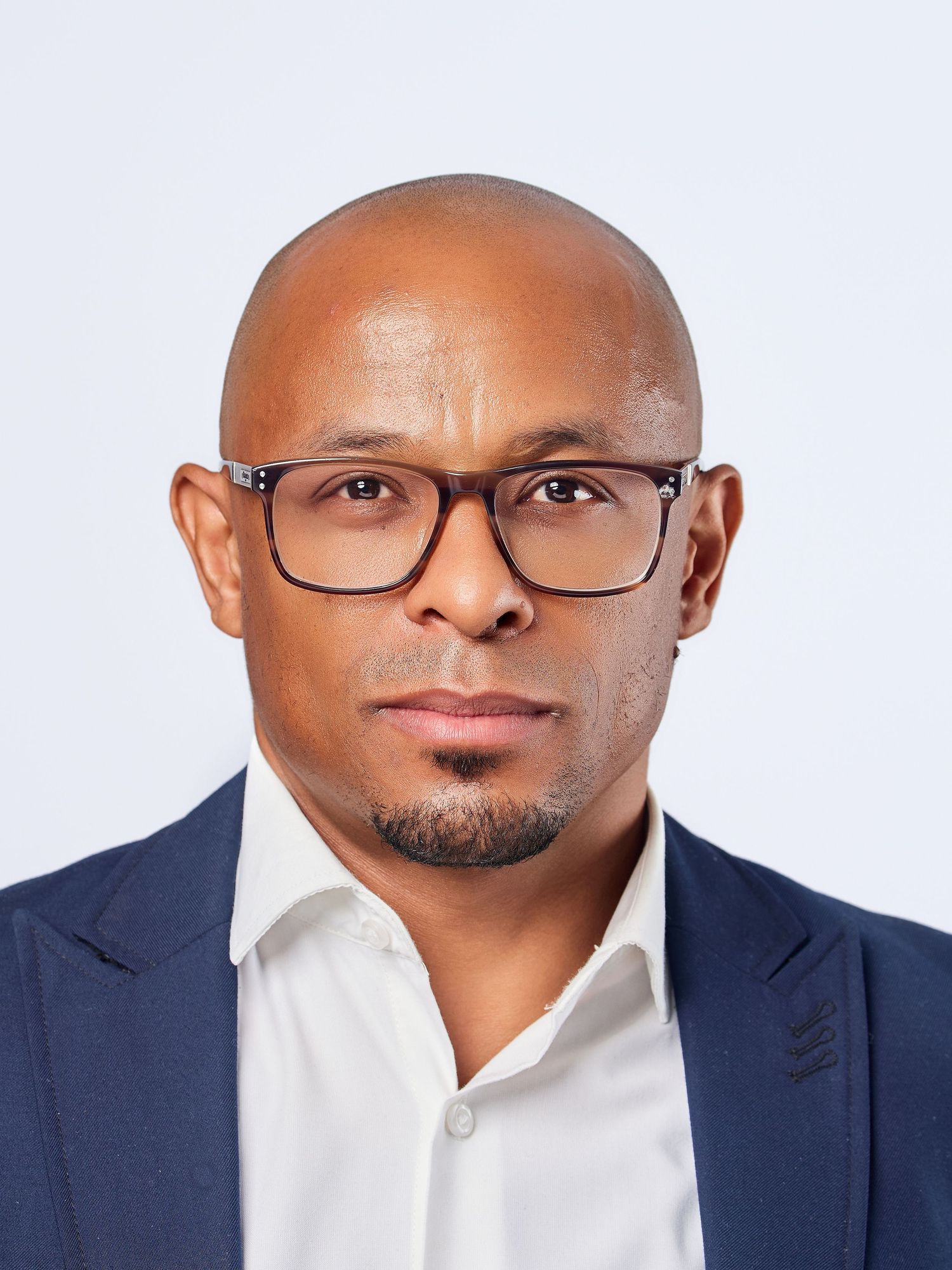 Bald Black man wearing rectangular glasses and navy blazer with white shirt against light gray background.