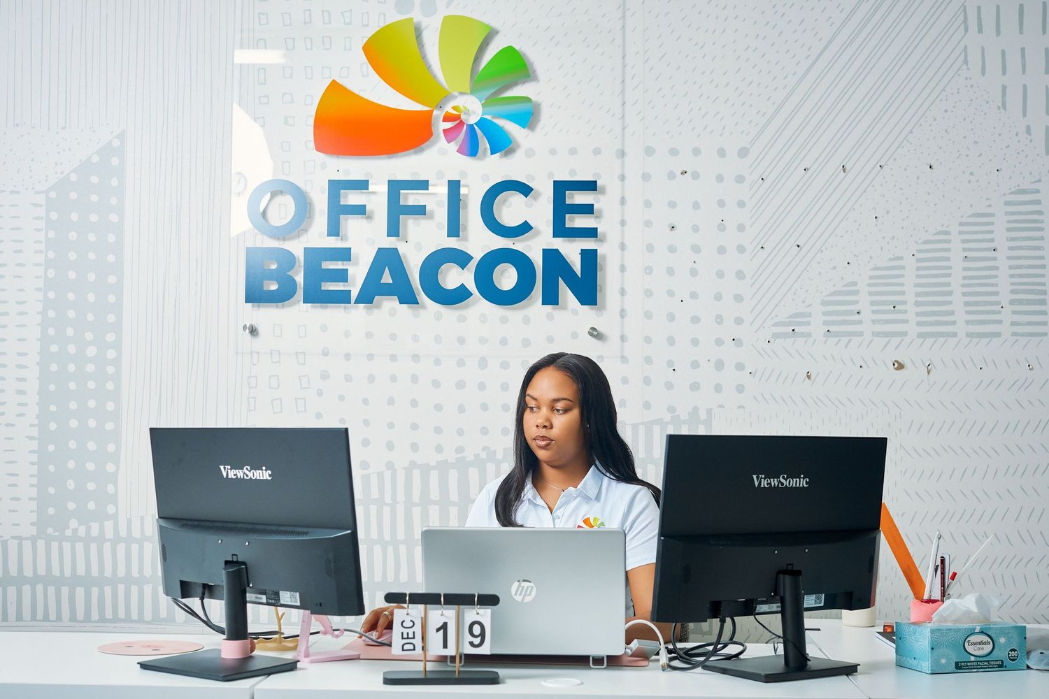 Young woman working at a reception desk with dual monitors at Office Beacon headquarters.