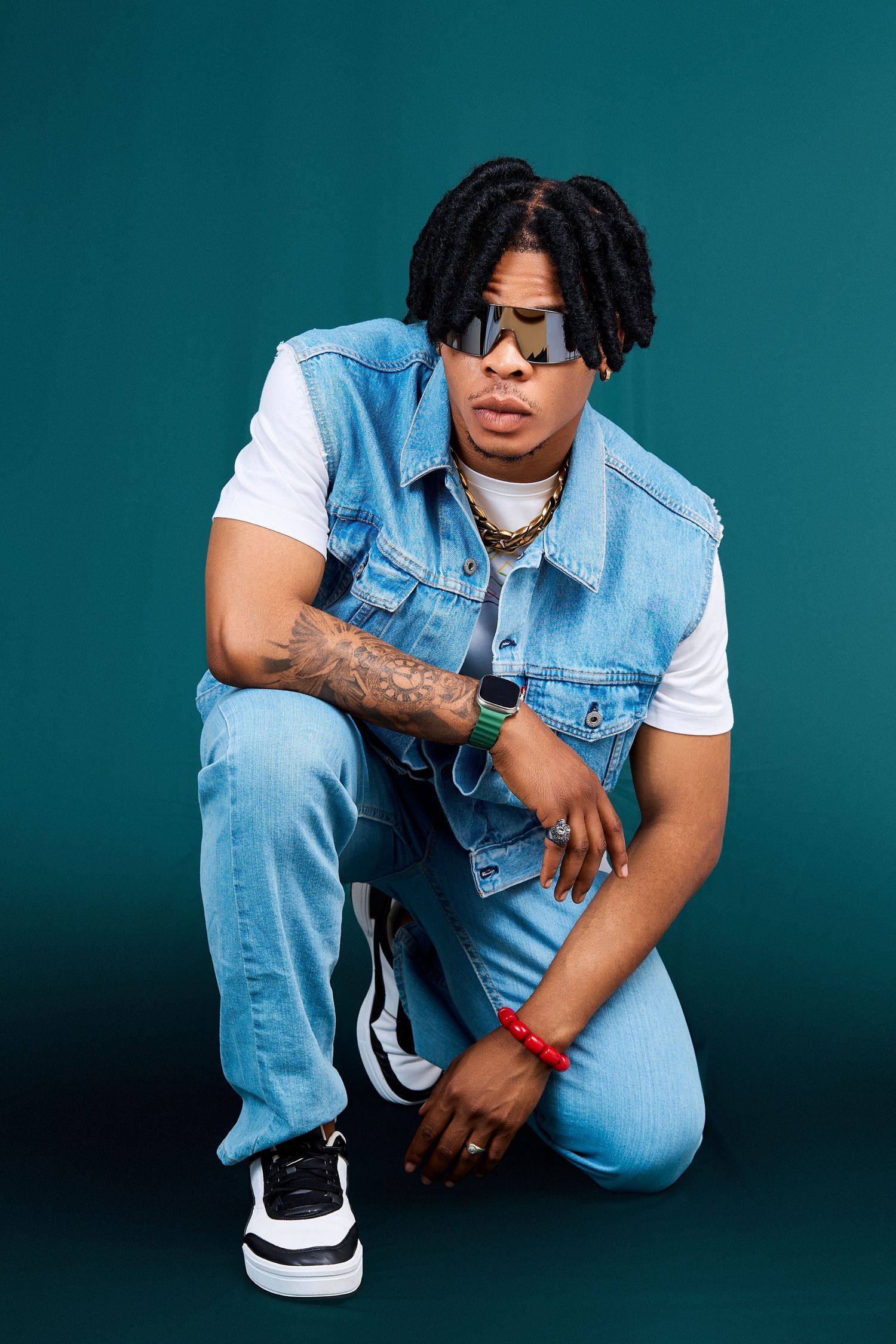 Fashionable Black man with dreadlocks in denim vest and jeans crouches on teal background wearing sunglasses.