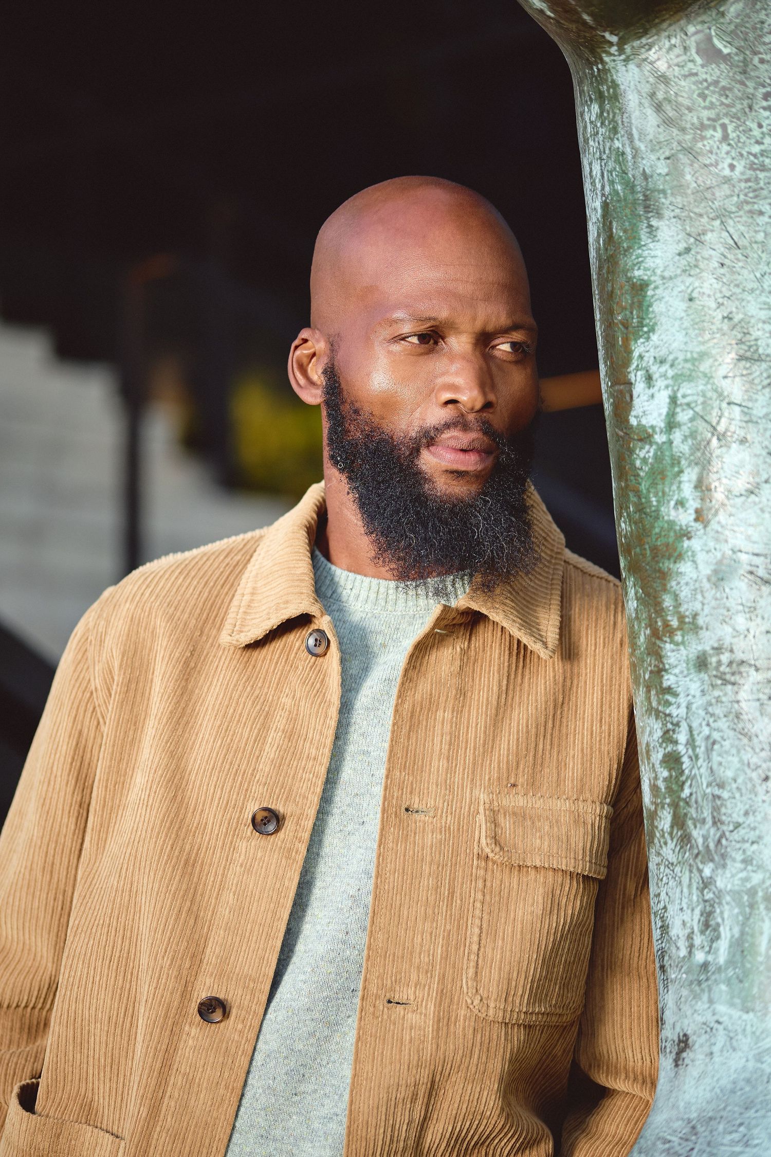 Bald man with full beard wearing tan corduroy jacket leans against weathered bronze statue outdoors.