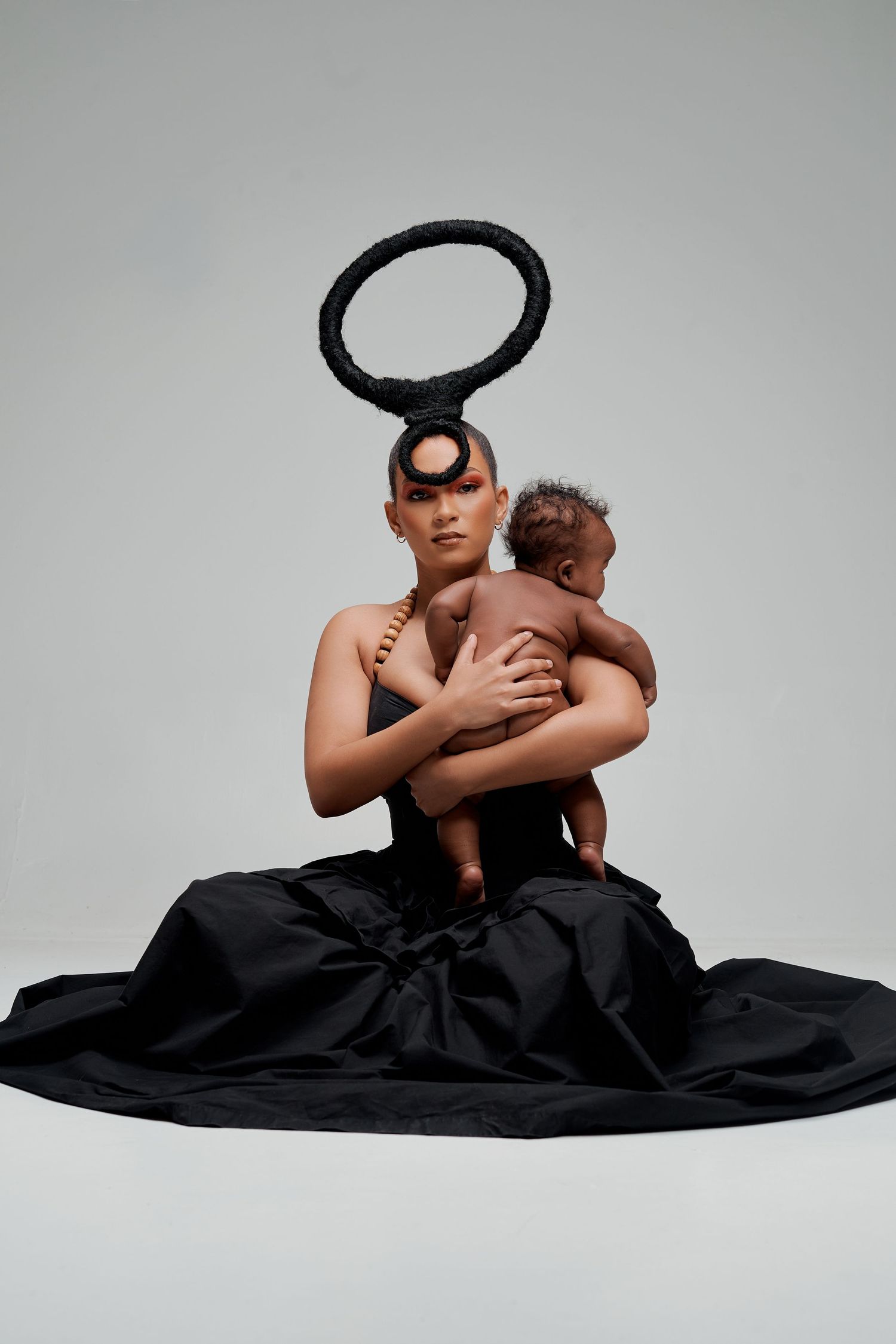 Woman in black gown with circular headpiece holds a baby in an artistic studio portrait.