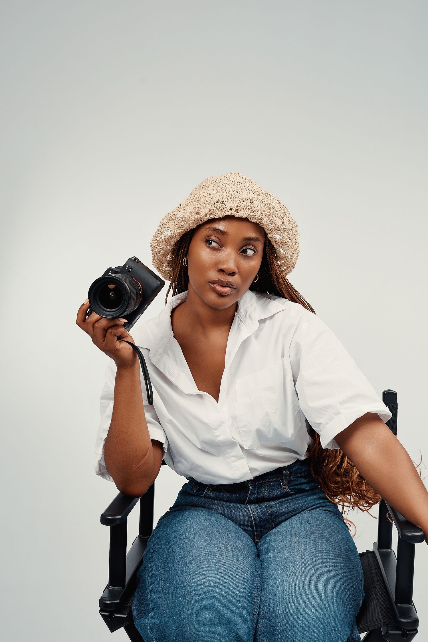 Black woman in straw hat and white shirt sits in director chair holding a camera against gray background.