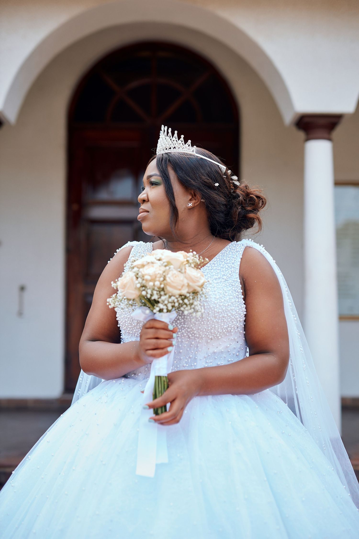 Stunning Black bride in white ball gown with veil and tiara holding bouquet outside church venue.