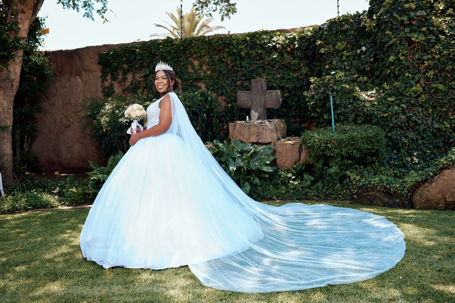 Bride in white ball gown with long train holding bouquet in garden near stone cross.