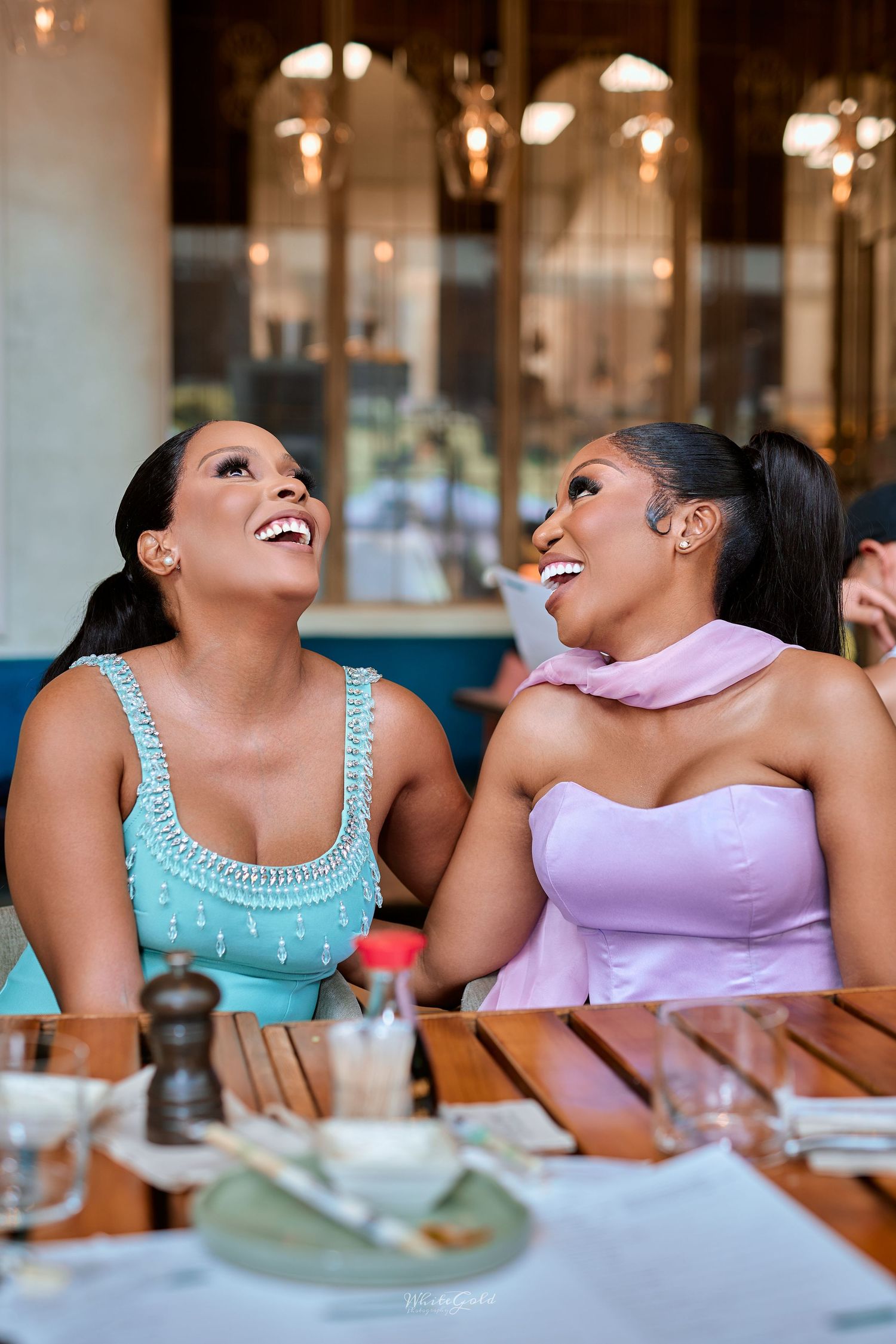 Two women in teal and pink outfits laugh together at a restaurant dining table.