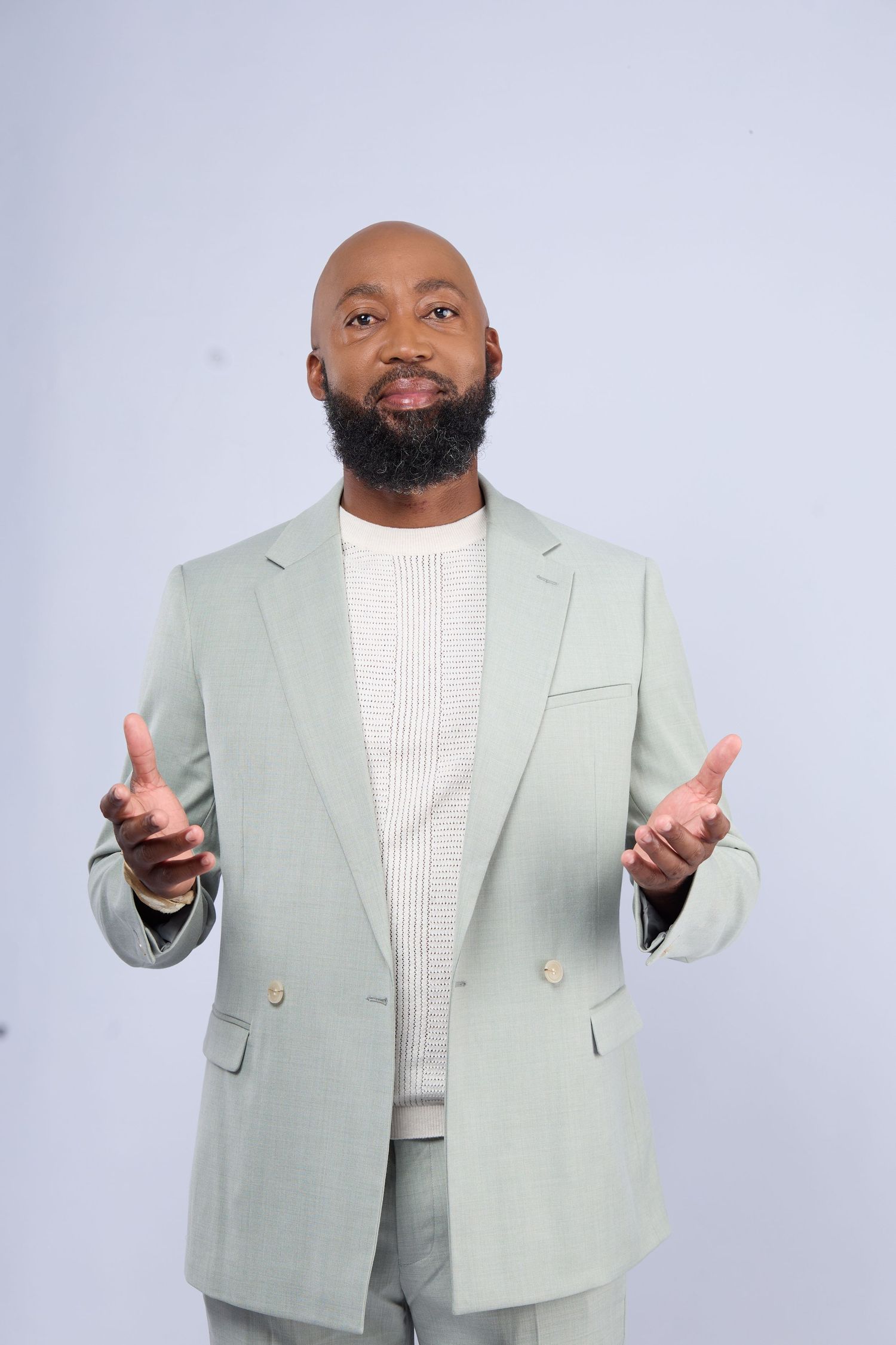 Bald Black man with beard wearing sage green double-breasted suit gestures with both hands against light gray background.