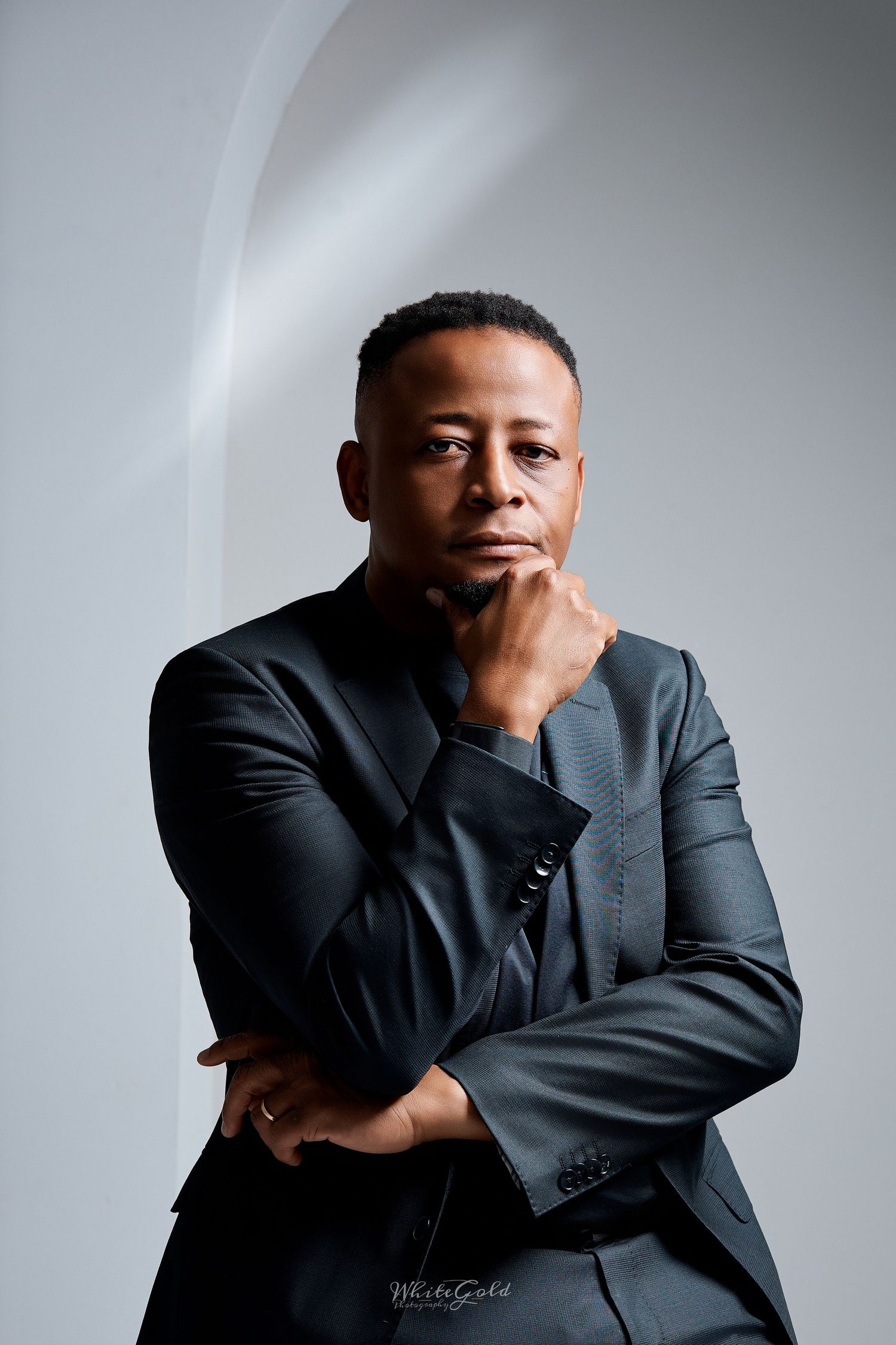 Distinguished Black man in dark suit with hand on chin in thoughtful pose leaning against white wall.