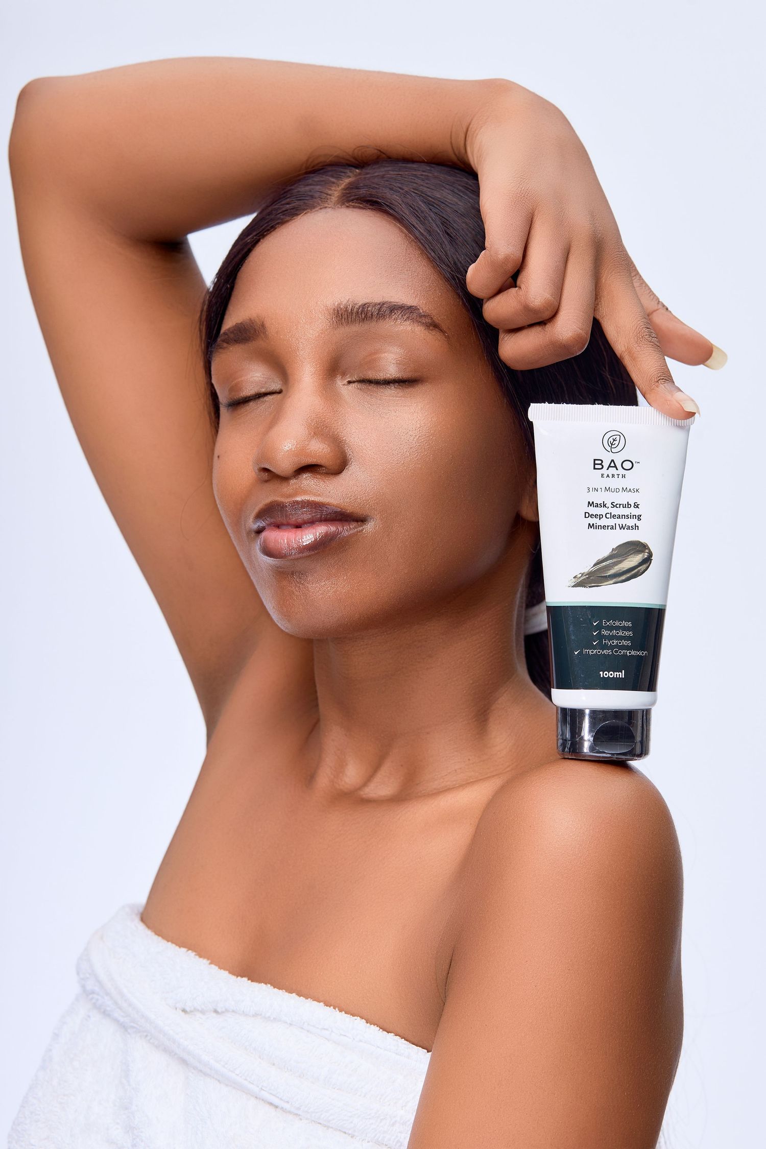 Woman wrapped in towel holding BAO skincare face wash tube with eyes closed on light background.