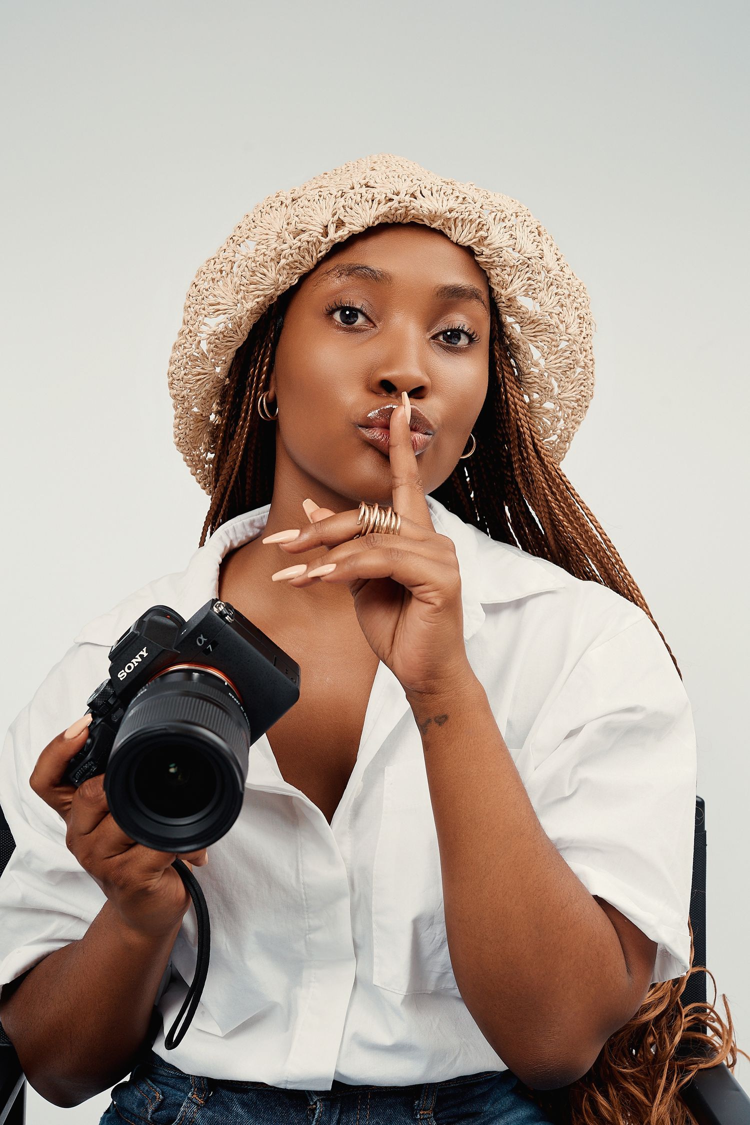 A Black woman in a straw hat holds a Sony camera and makes a shush gesture in a studio.