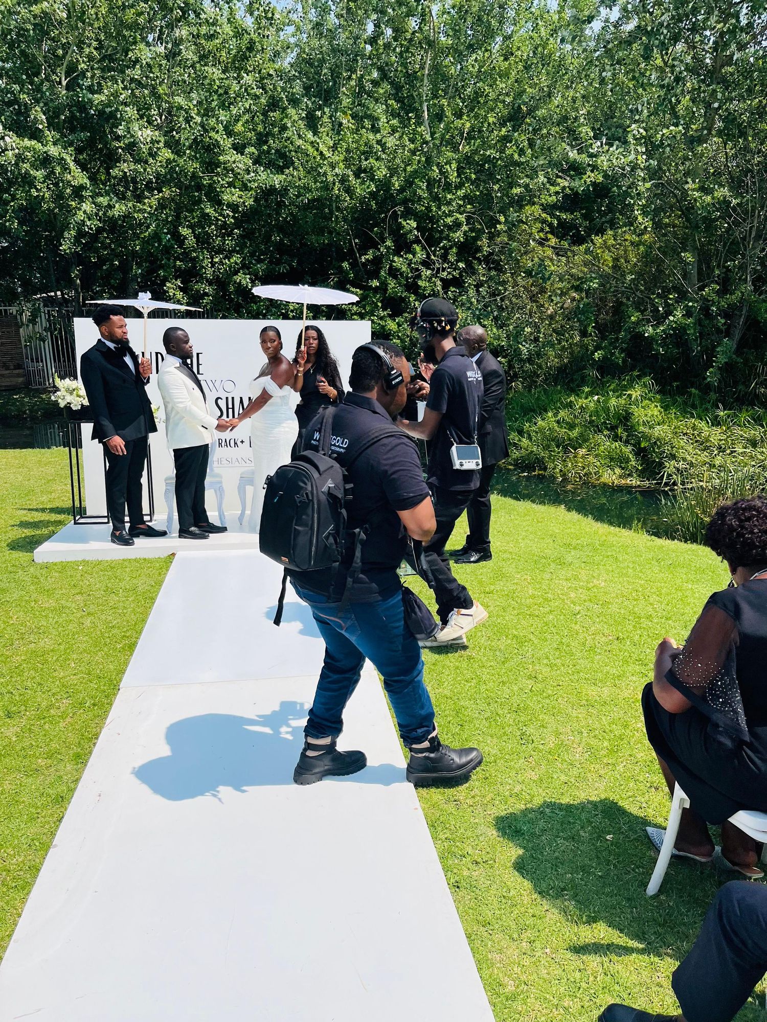 Outdoor wedding ceremony on green lawn with wedding party at altar and photographers capturing the moment.