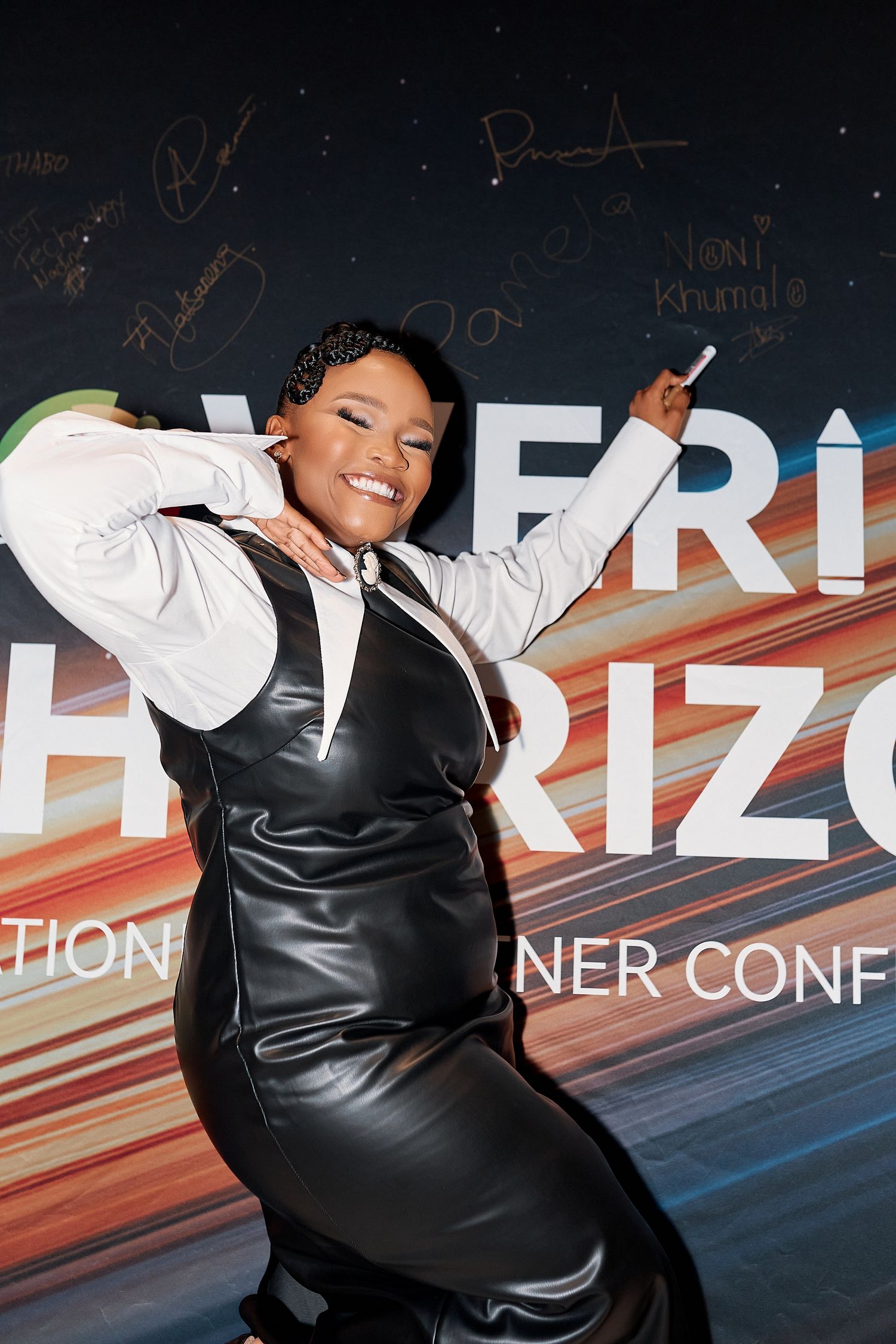 A smiling woman in a black leather dress poses confidently at the New Horizons Innovation Partner Conference backdrop.