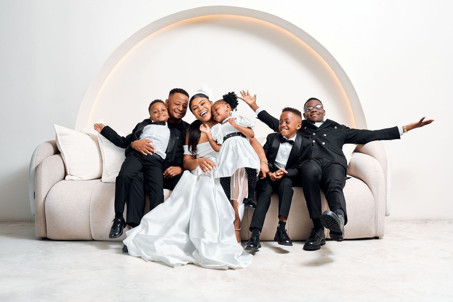 Joyful Black family in formal black and white attire celebrating together on a cream sofa with arch backdrop.