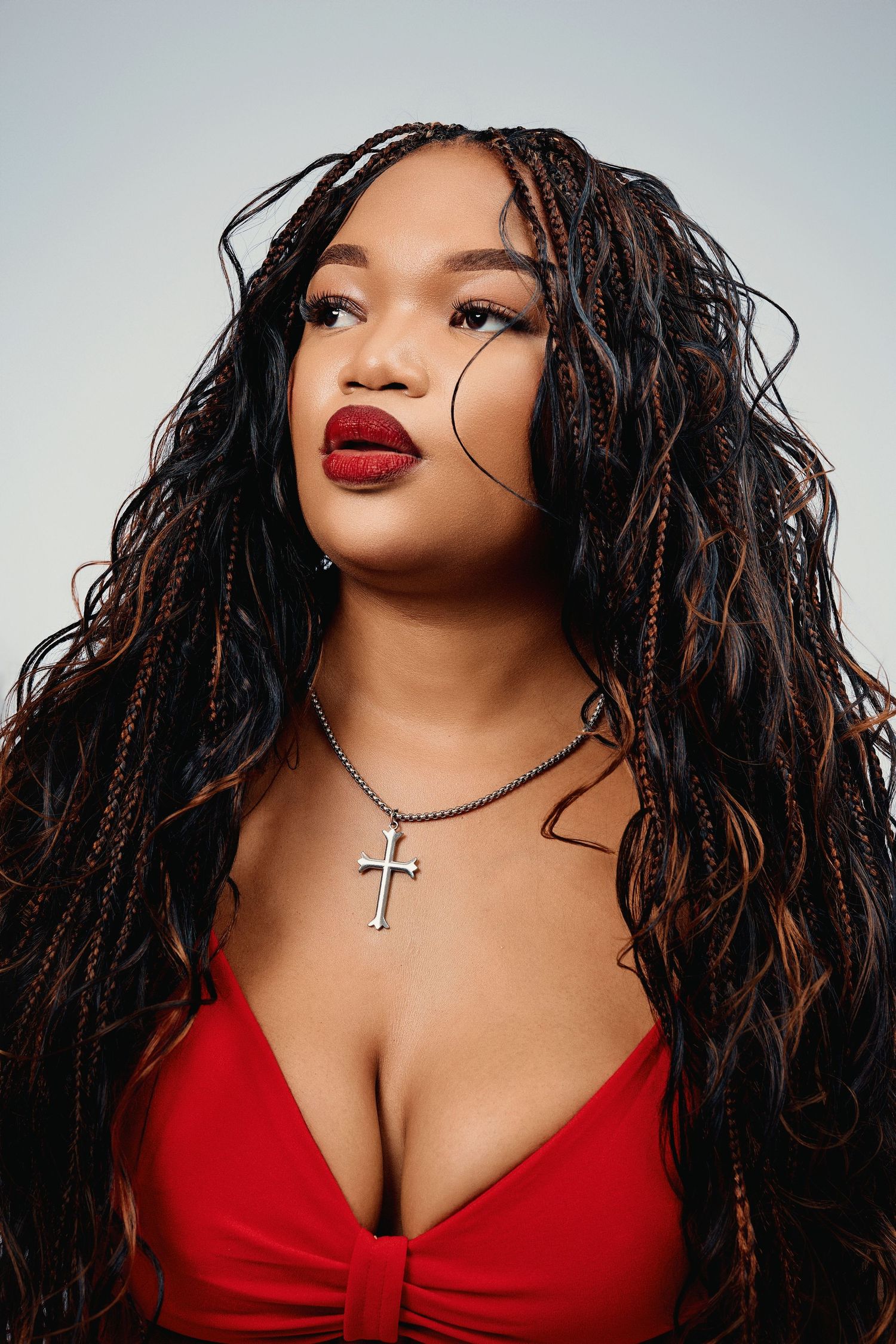 A Black woman with long box braids and red lips wears a red top and cross necklace in a studio portrait.