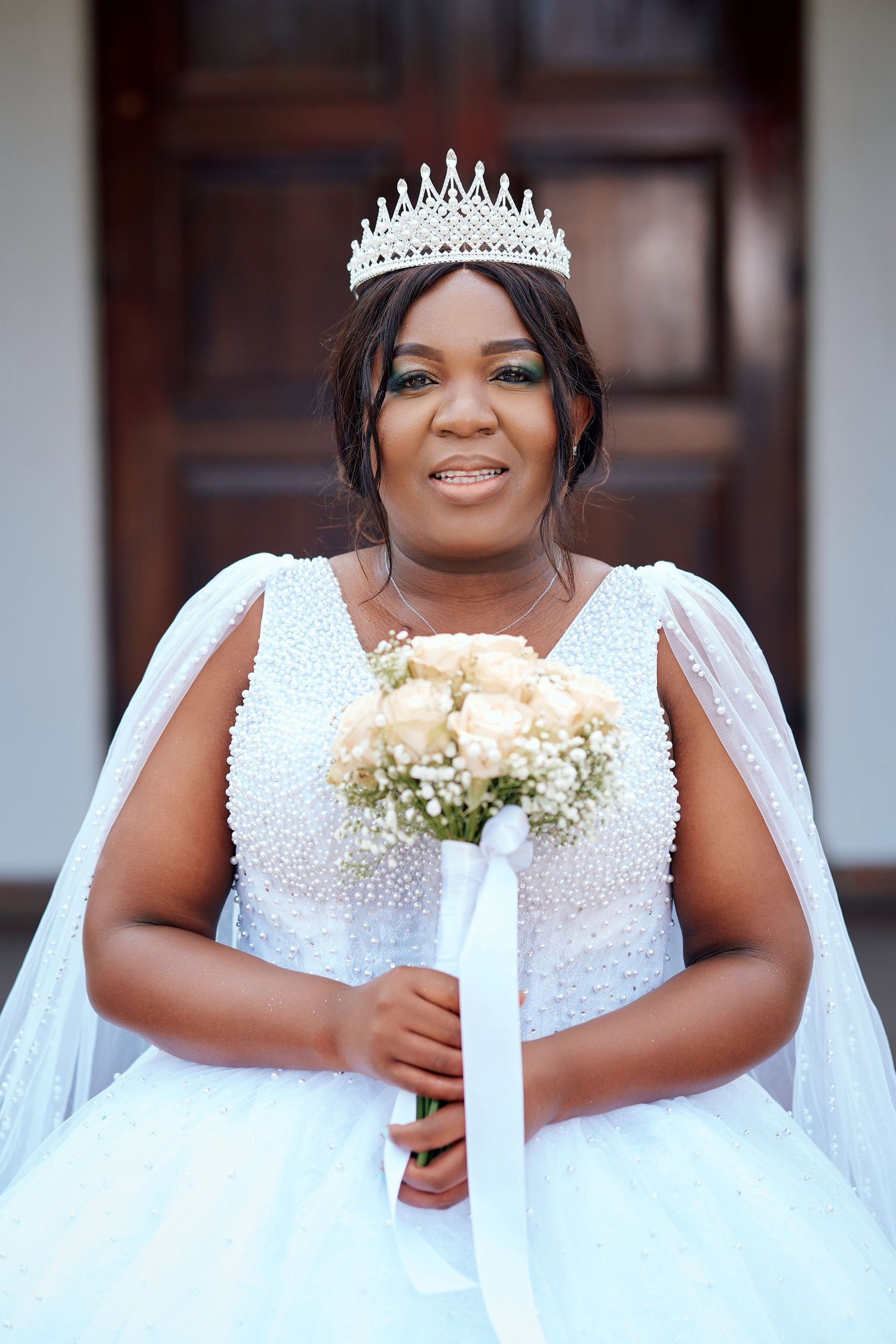 African American bride in pearl-embellished white gown and diamond tiara holds rose bouquet in front of wooden doors.