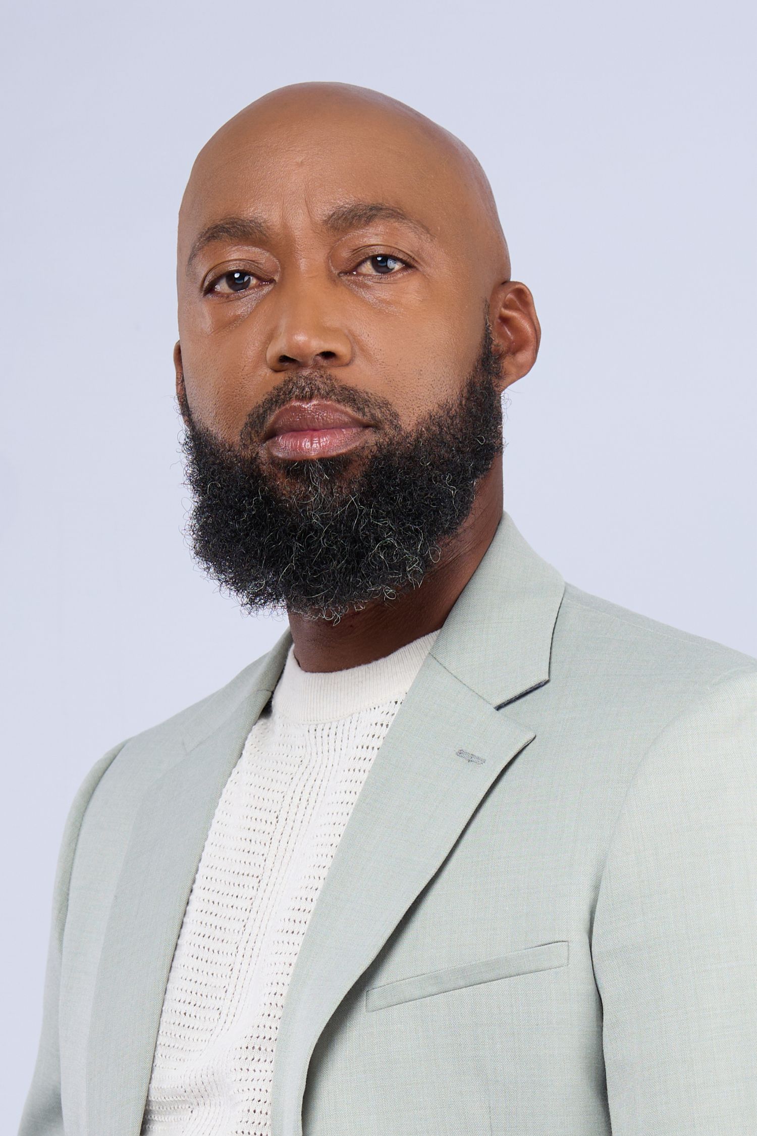 Bald Black man with full beard wearing a light sage blazer over white sweater against gray background.