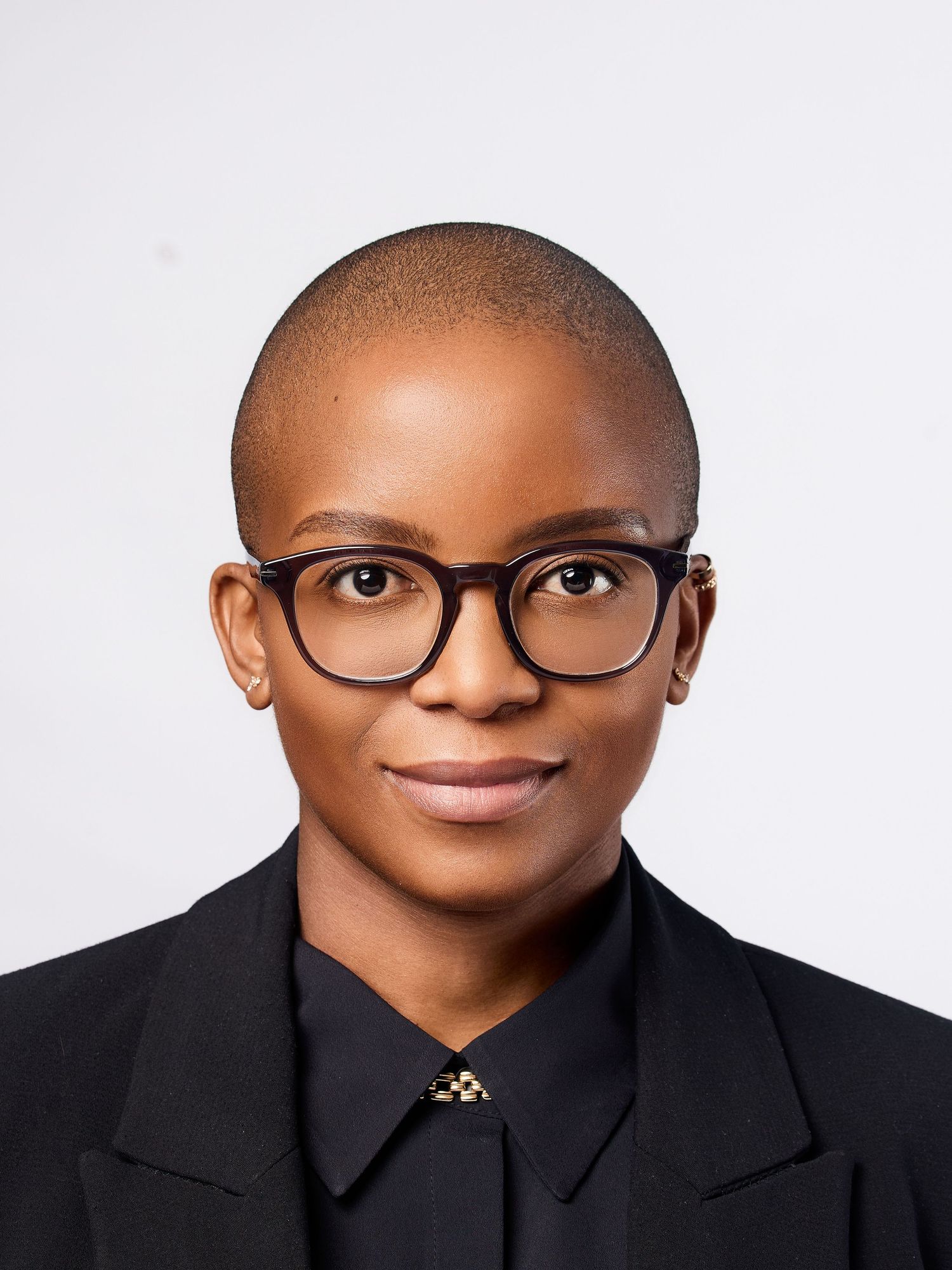 Professional headshot of a woman with a shaved head wearing black-rimmed glasses and a black blazer.