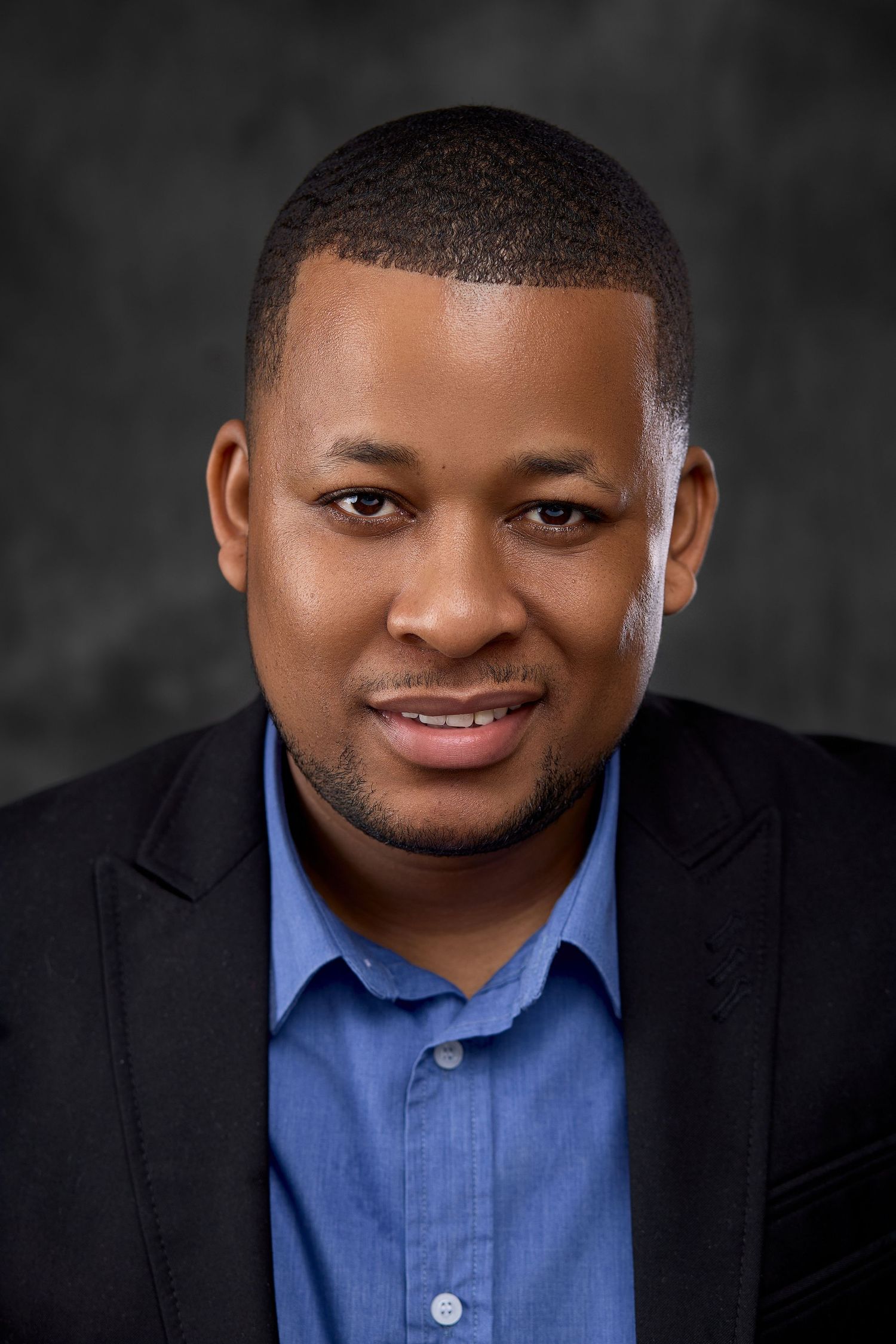 Professional headshot of a smiling Black man in a blue shirt and black blazer against a dark background.