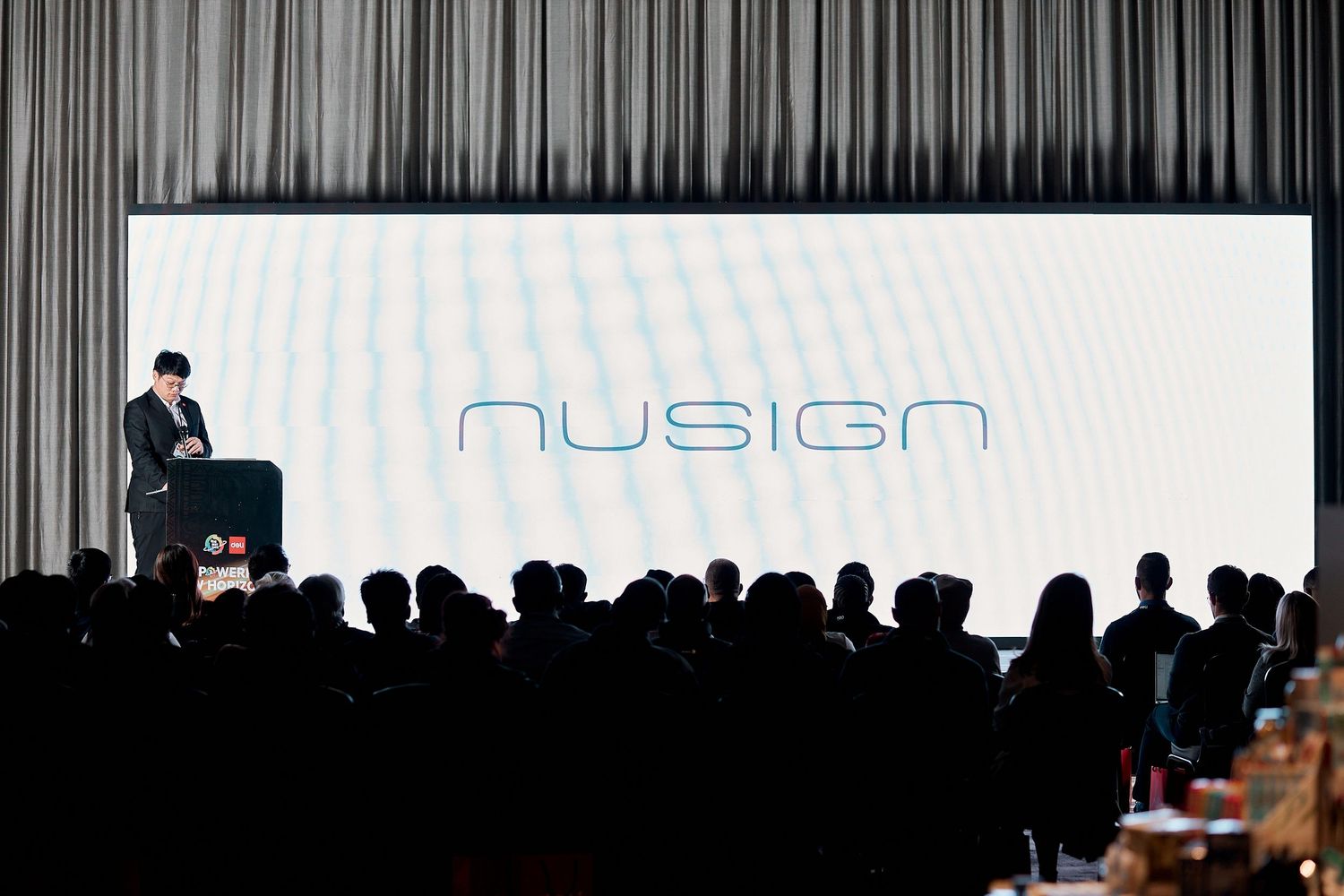 A presenter on stage at a Nusign brand event, speaking to a large audience in a darkened conference hall.