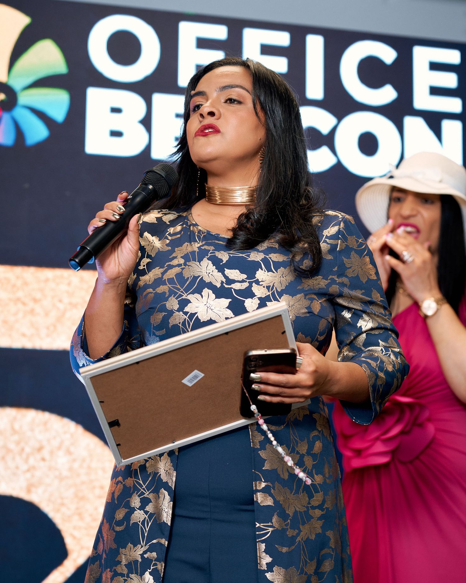 Woman in blue floral dress speaking into microphone at Office Beacon event holding framed award.
